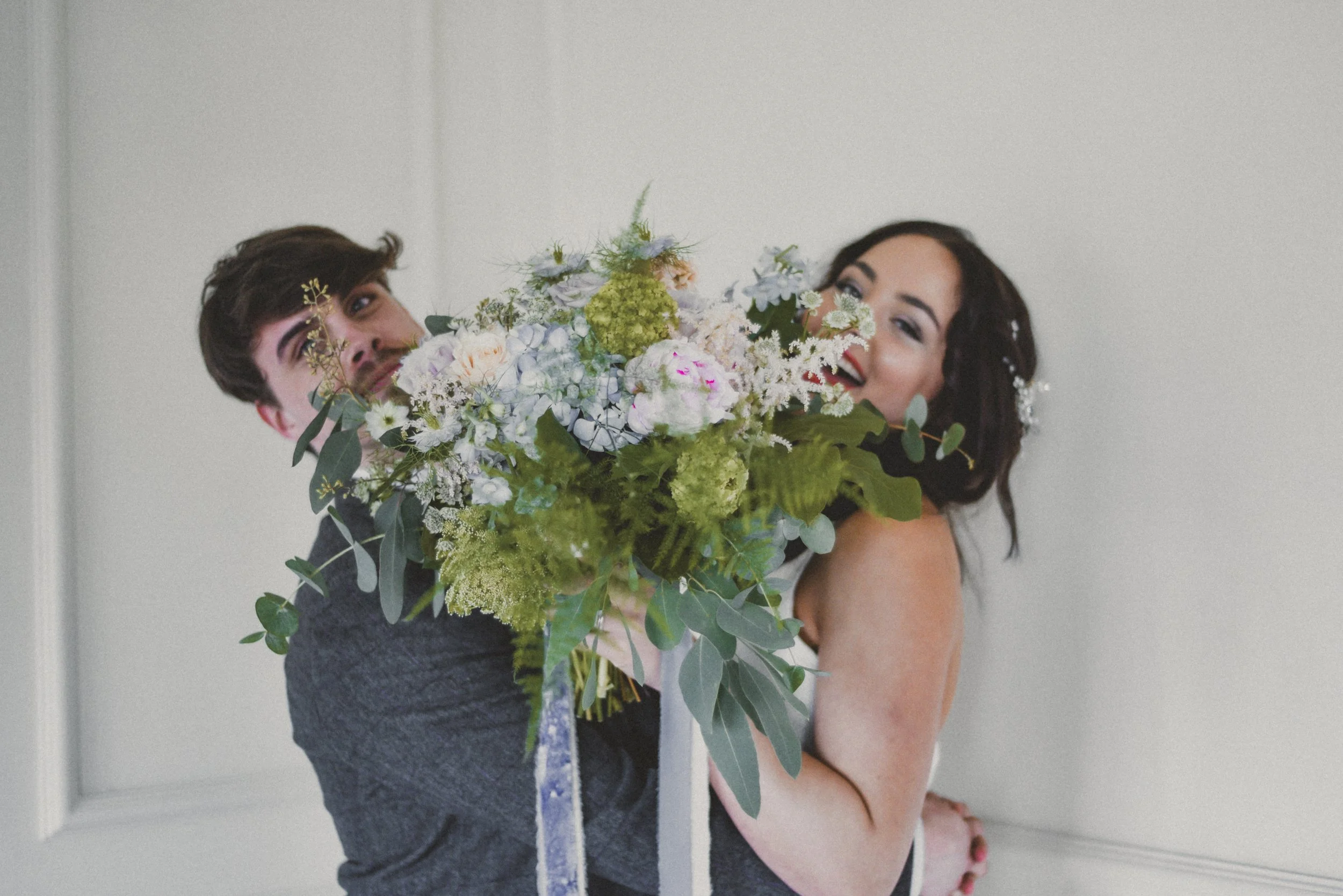 A couple embracing, holding a large bouquet of flowers, with the woman smiling and the man looking at the camera, against a plain light-colored background.