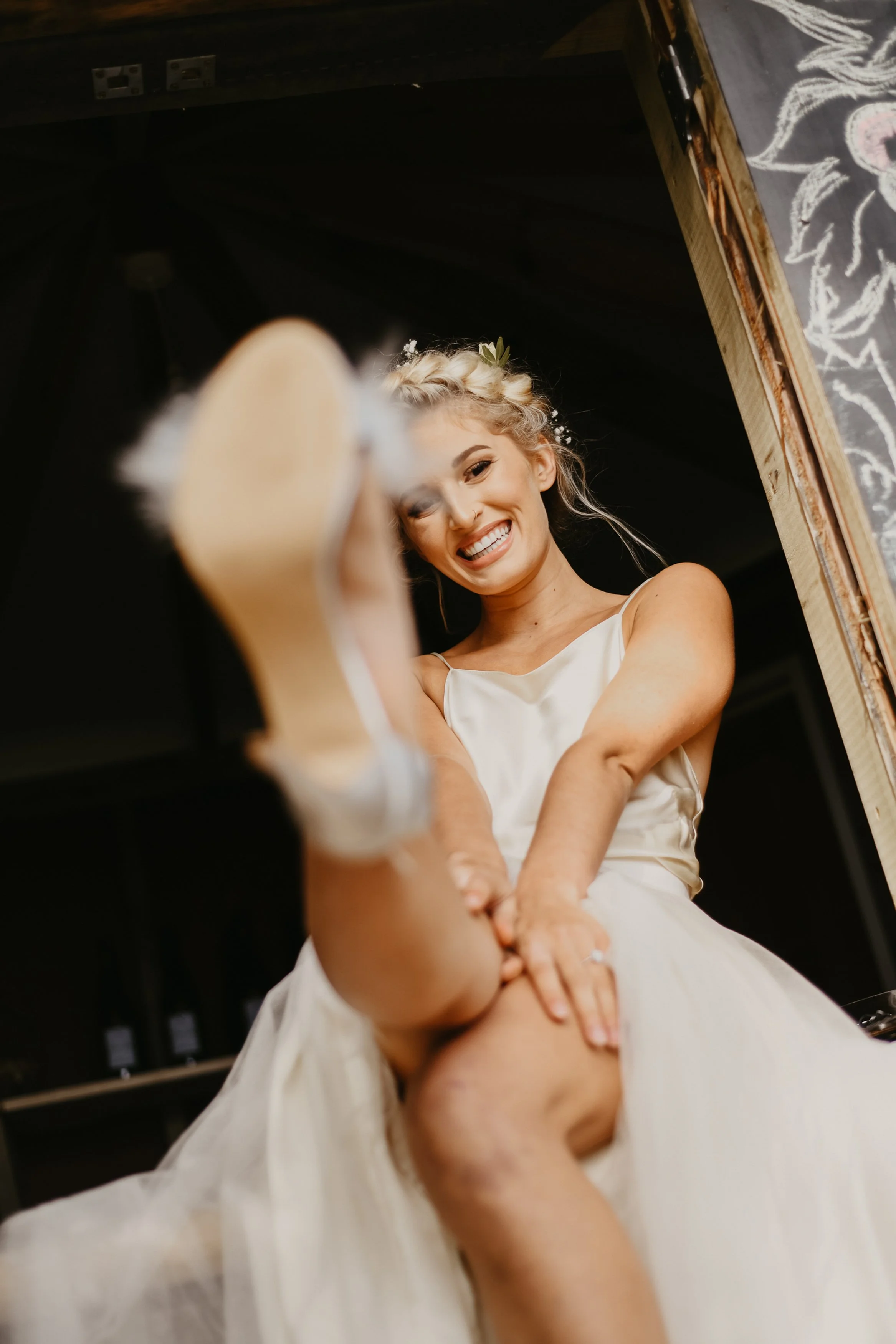 Brunette woman in a white dress smiling while putting on her shoe, with a floral crown, sitting in a rustic setting.