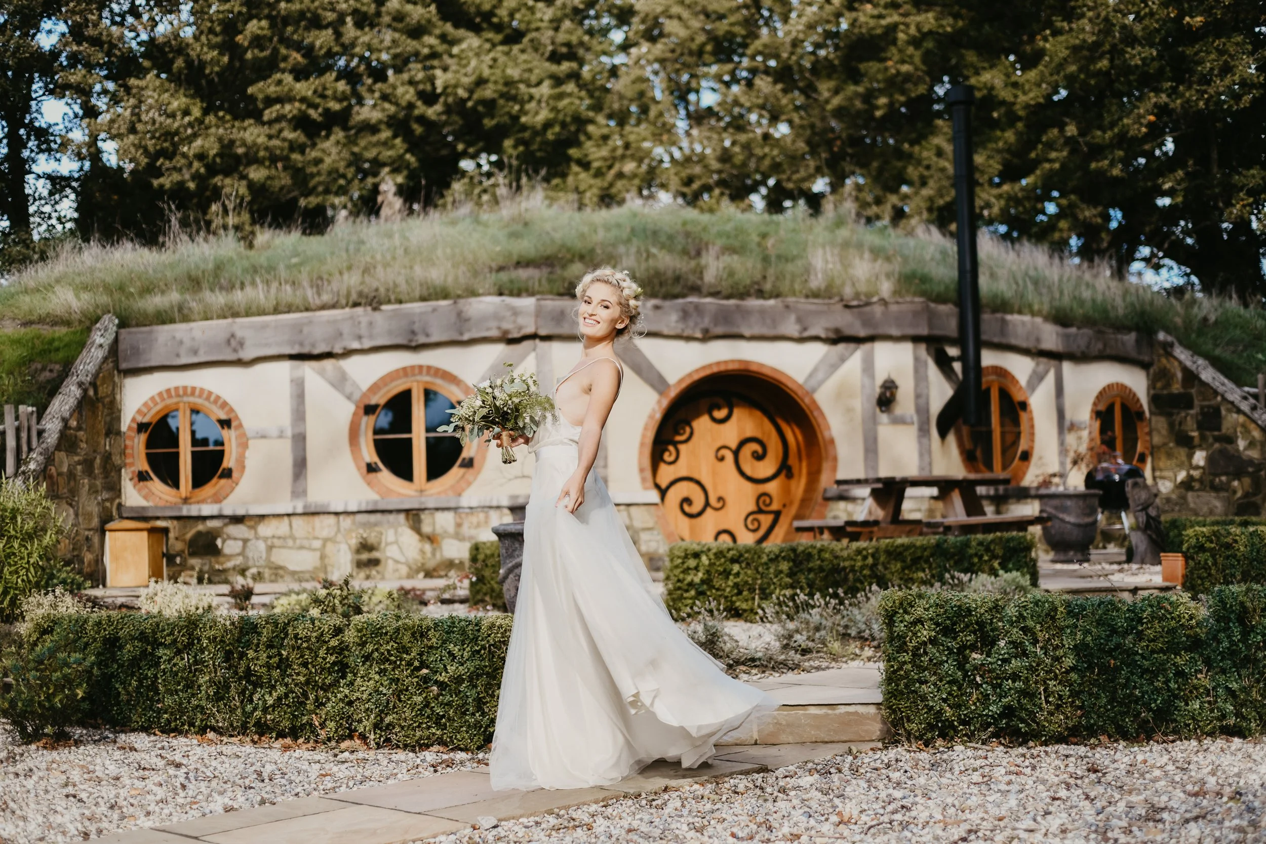 A bride standing outdoors in front of a small house with a rounded door and circular windows, holding a bouquet, smiling, in a white wedding dress. A Hobbit style house at a luxury wedding destination.