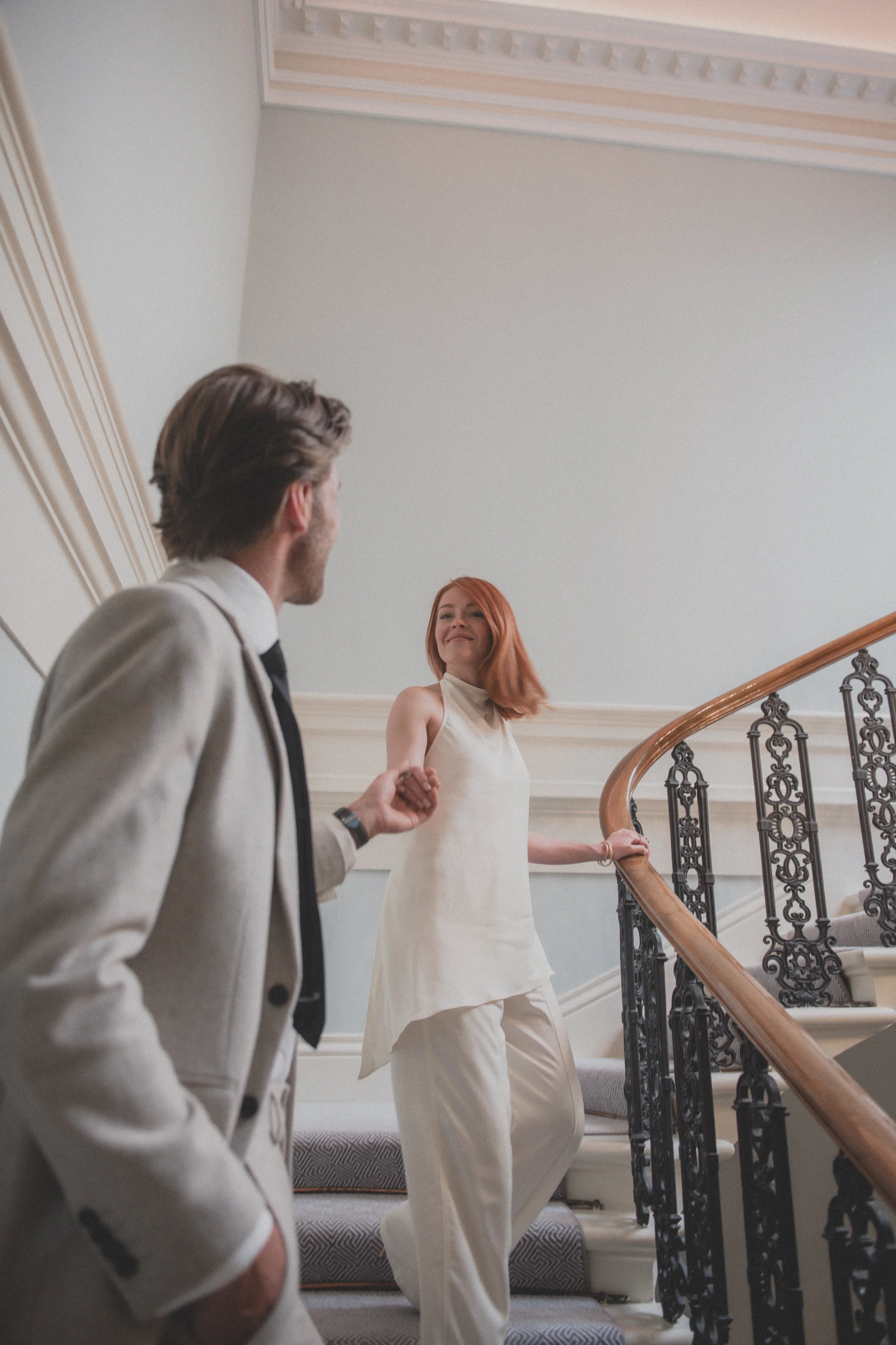 A man in a light-colored suit looks at a woman with red hair dressed in a white sleeveless outfit, standing on a curved staircase with an ornate black iron railing and wooden handrail.