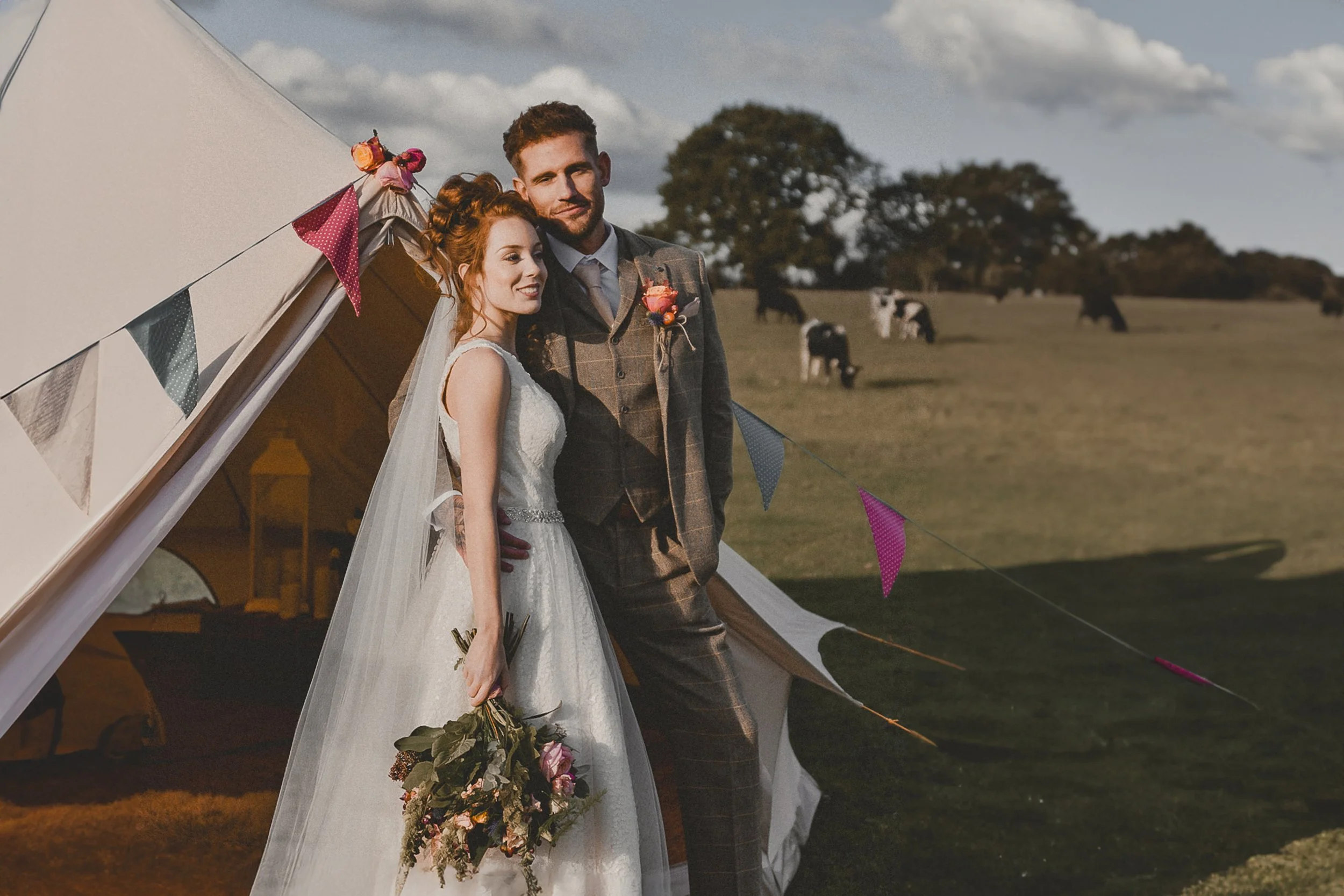 A bride and groom standing outside in front of a white tent decorated with colorful bunting flags. The bride is holding a bouquet of flowers, and the groom has a boutonniere. They are smiling with their arms around each other, with a scenic landscape
