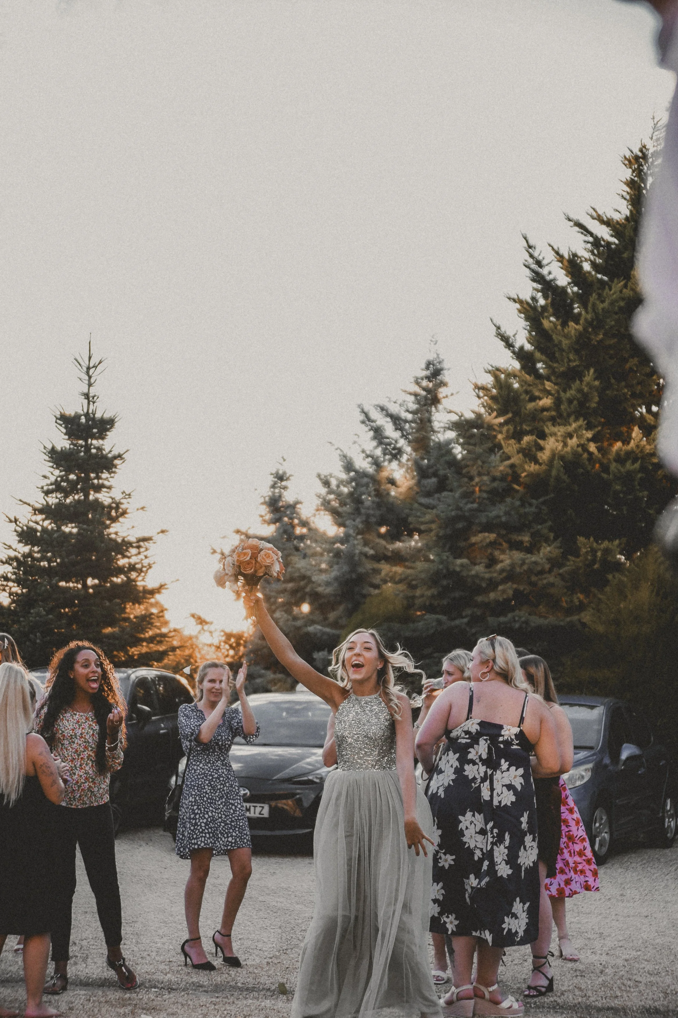 A group of women celebrating outdoors during sunset, with one woman in a silver and nude gown holding a bouquet of roses in the air, surrounded by others clapping and cheering, against a backdrop of tall trees and parked cars.