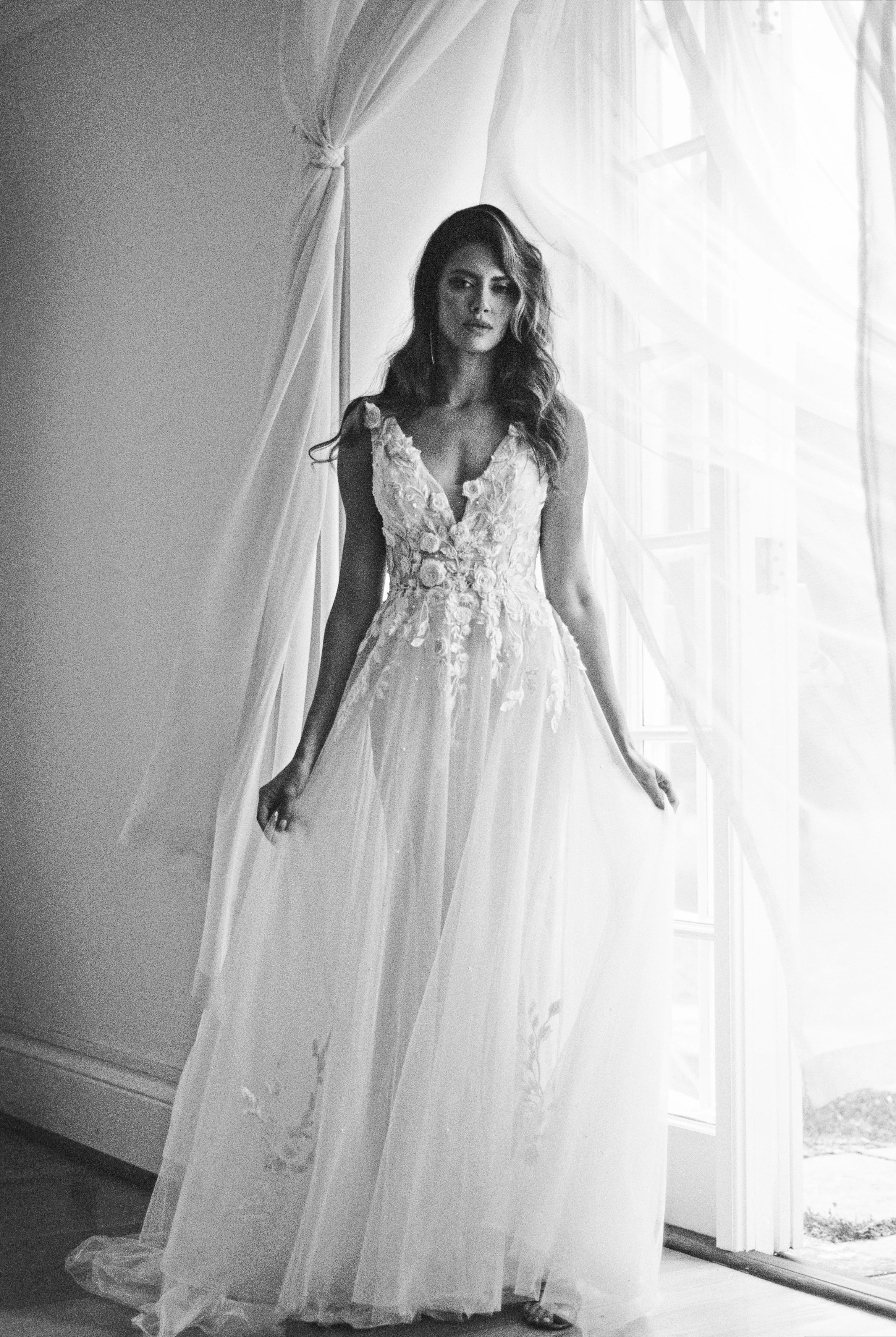 A woman in a bridal gown standing near a window with sheer curtains, black and white photo.