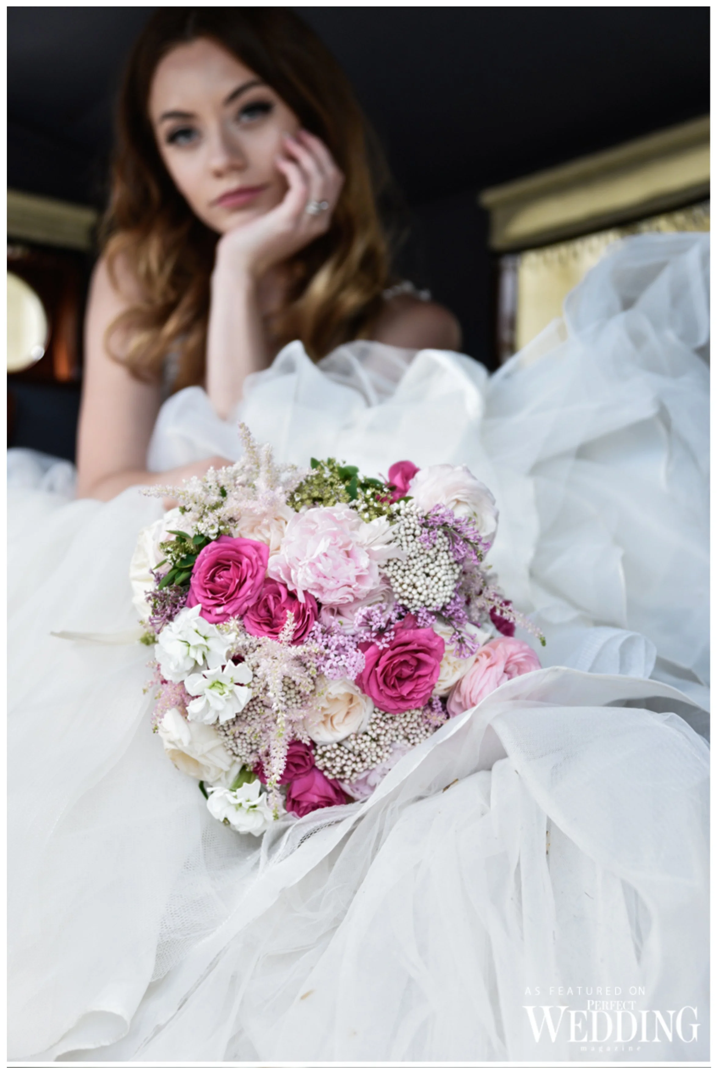 A woman in a wedding dress resting her head on her hand, with a bouquet of pink and white flowers in front of her, inside a room with dark walls and yellow curtains.