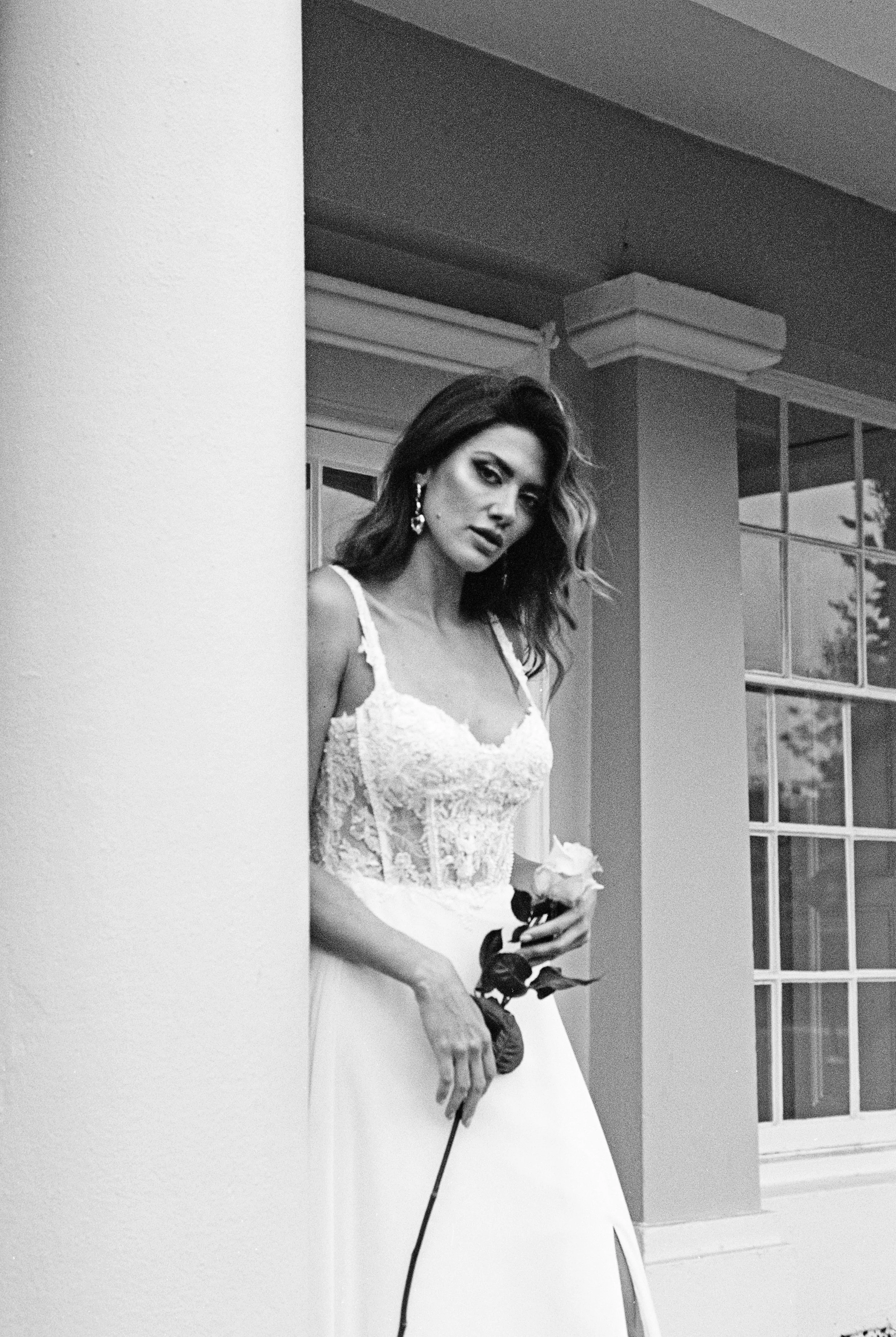 A woman in a wedding dress holding a rose, standing near a wall with a window in the background.