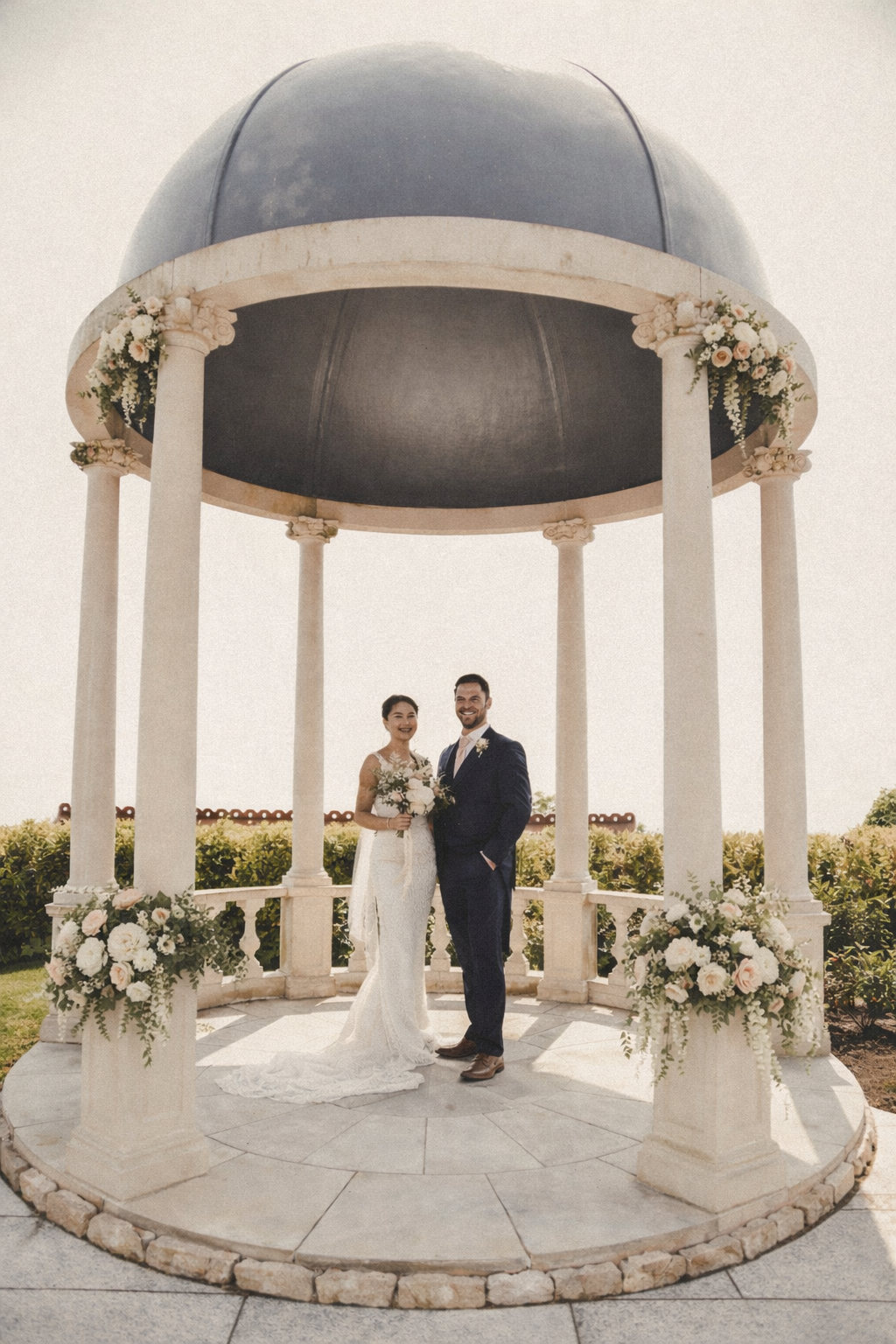 A newlywed couple standing under a decorative gazebo with floral arrangements, smiling at the camera.