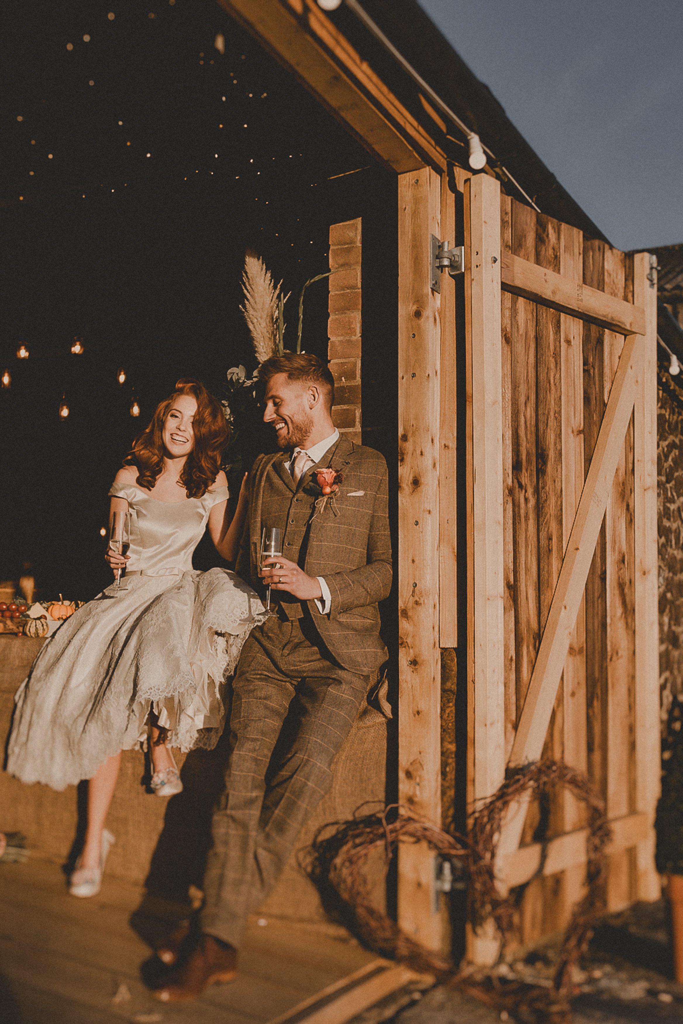 A bride and groom, dressed in wedding attire, sitting on a rustic wooden ledge inside a barn, holding champagne glasses and smiling at each other during their wedding celebration at night.