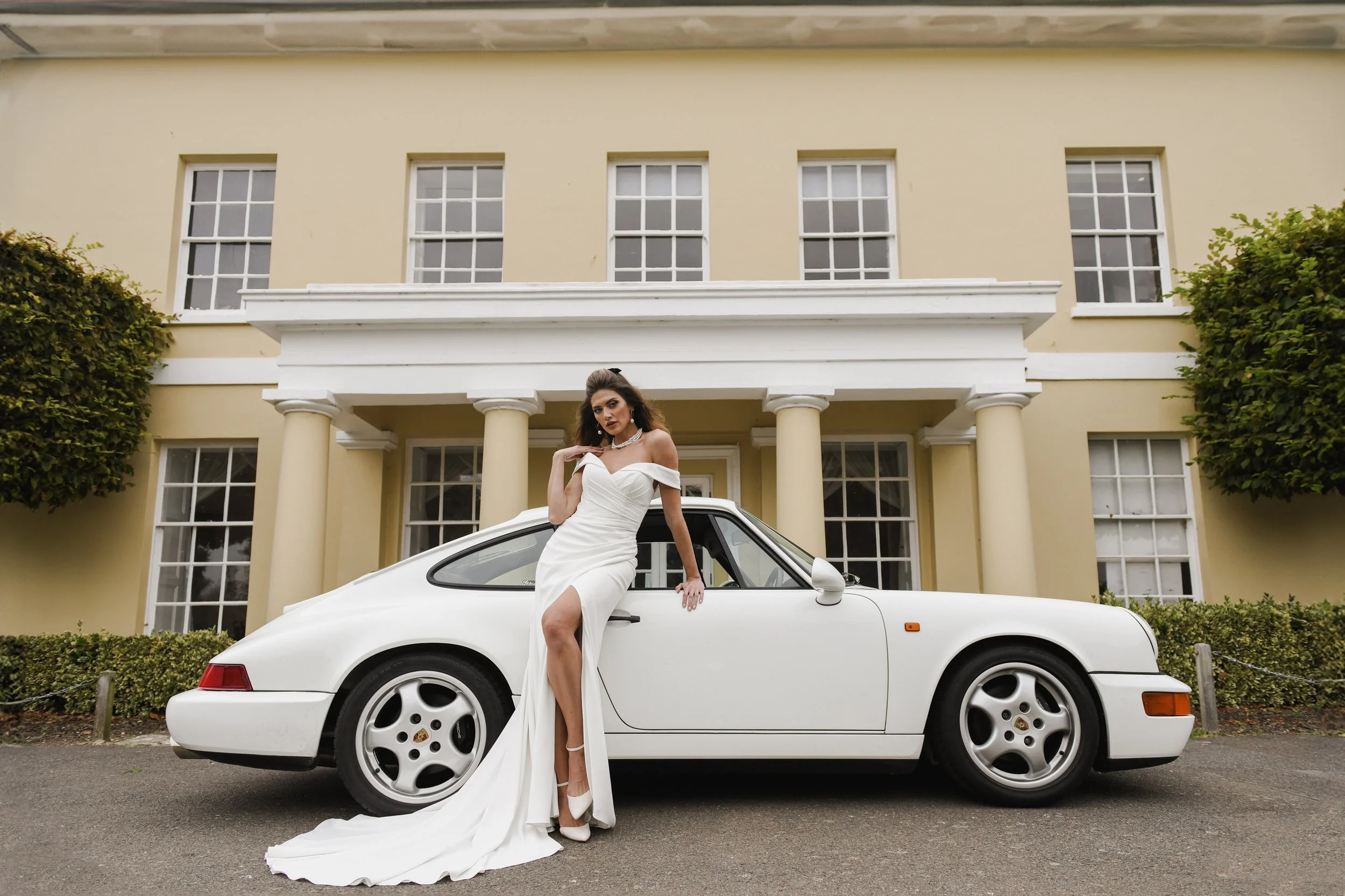 Woman in a white wedding dress posing next to a white vintage Porsche car in front of a yellow mansion with large windows and columns.