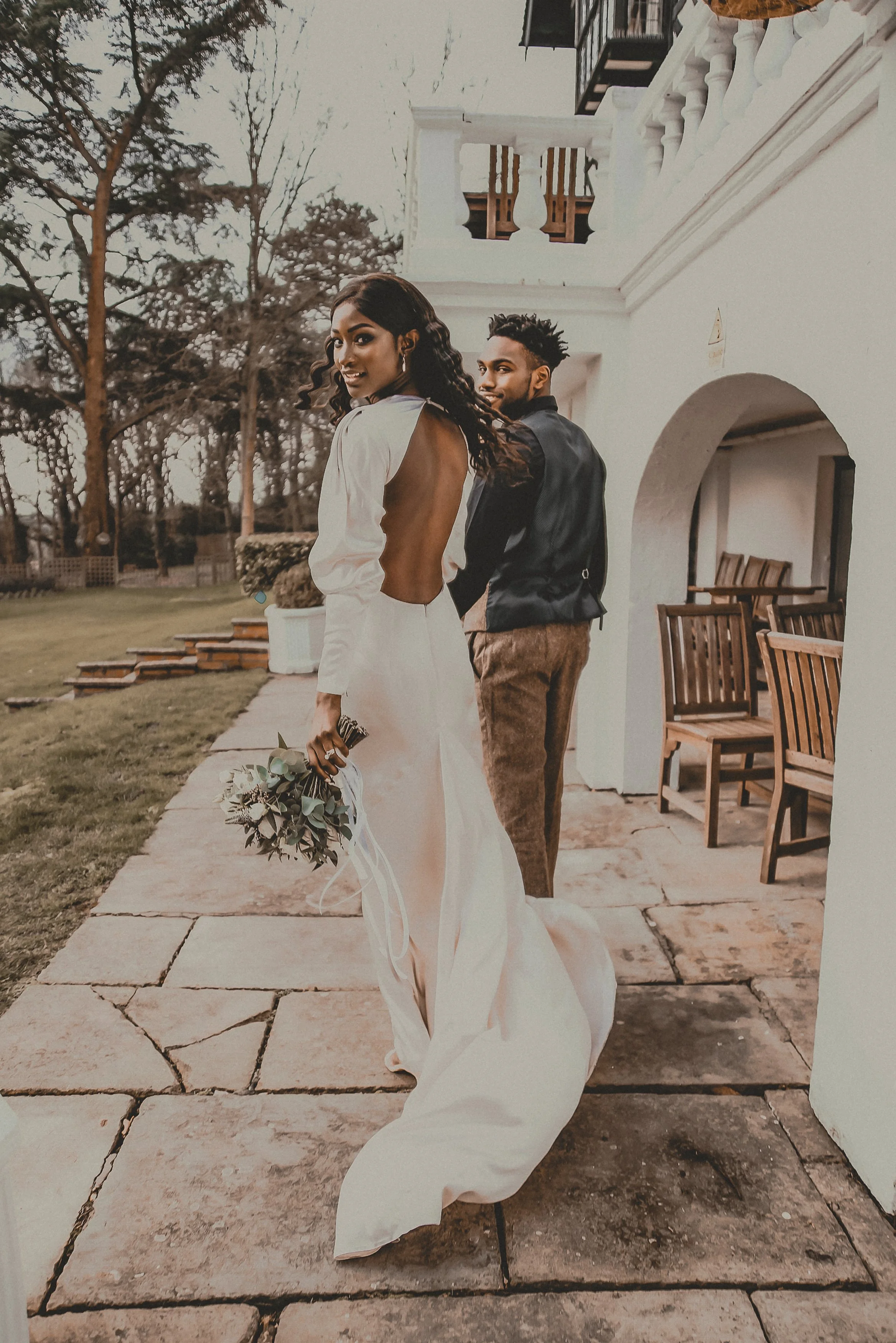 Bride in a backless wedding dress holding a bouquet standing on a stone pathway, with a groom in a dark jacket and brown pants behind her, outdoors near a white building with outdoor seating.