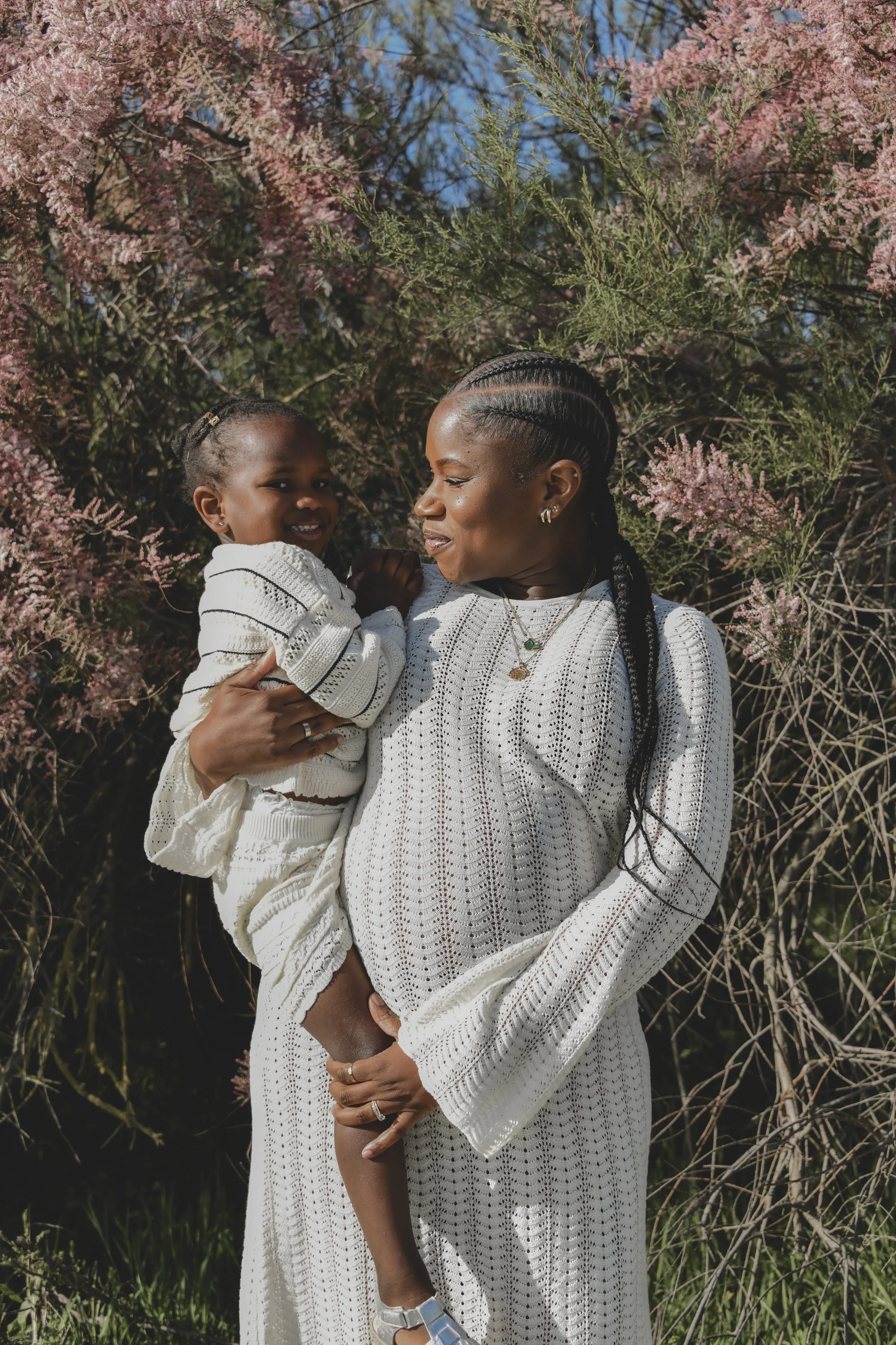 A woman holding a young girl in front of pink flowering bushes on a bright day.