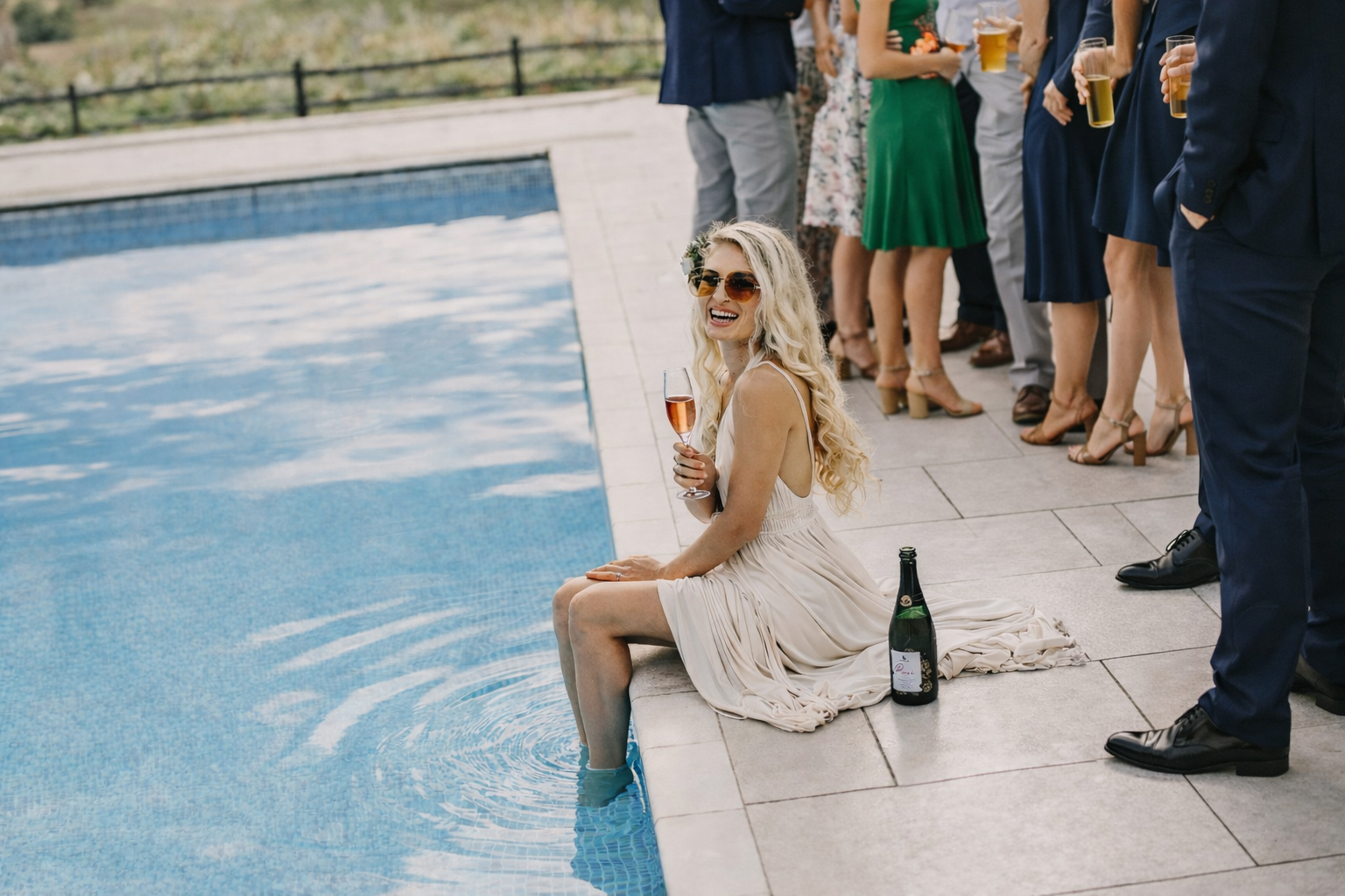A woman in a beige dress sitting by the edge of a pool with her feet in the water, holding a glass of rosé, surrounded by guests at a gathering, with a bottle of champagne nearby.