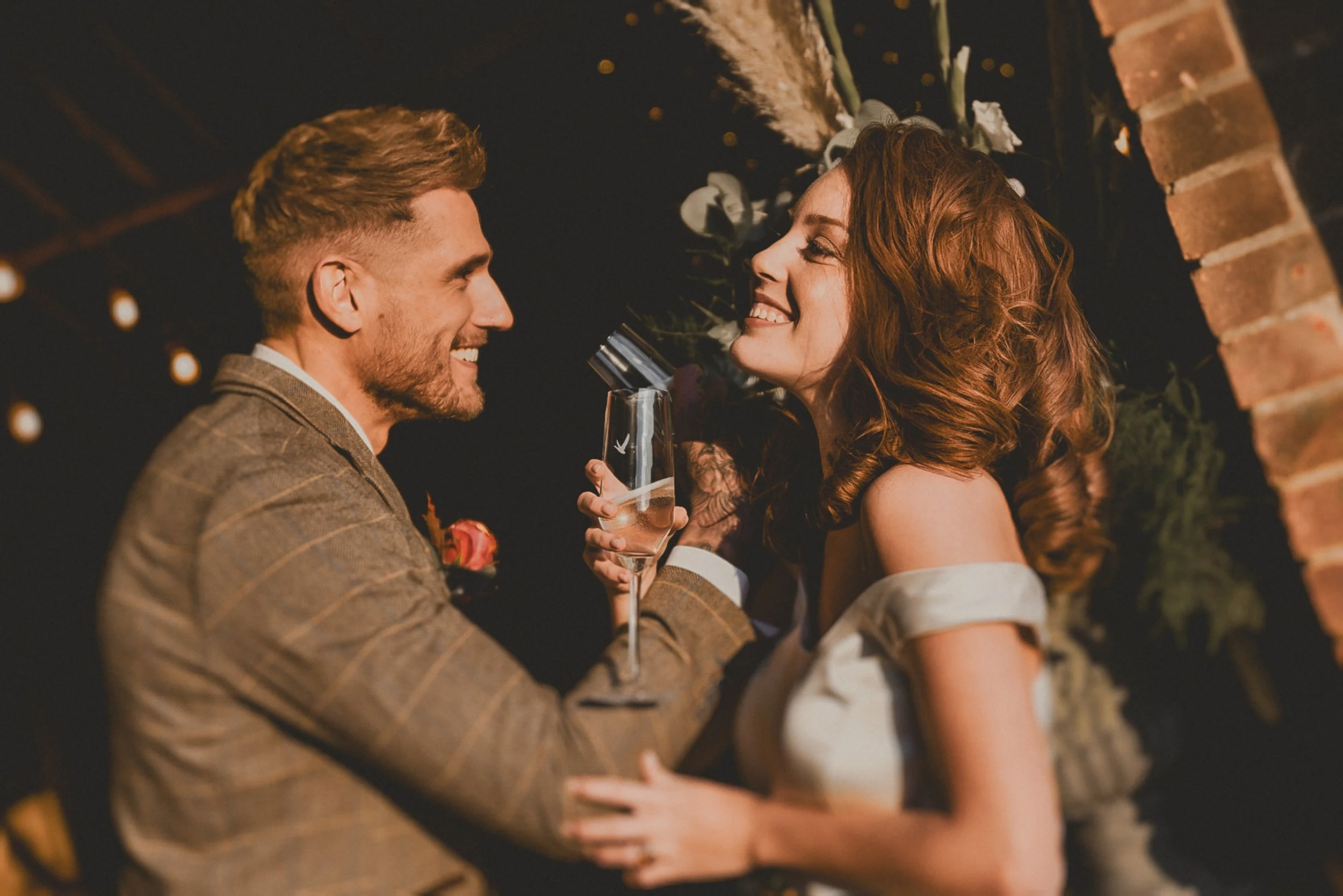A couple at a wedding celebration holding champagne glasses, smiling and looking at each other, surrounded by warm lighting and festive decorations.