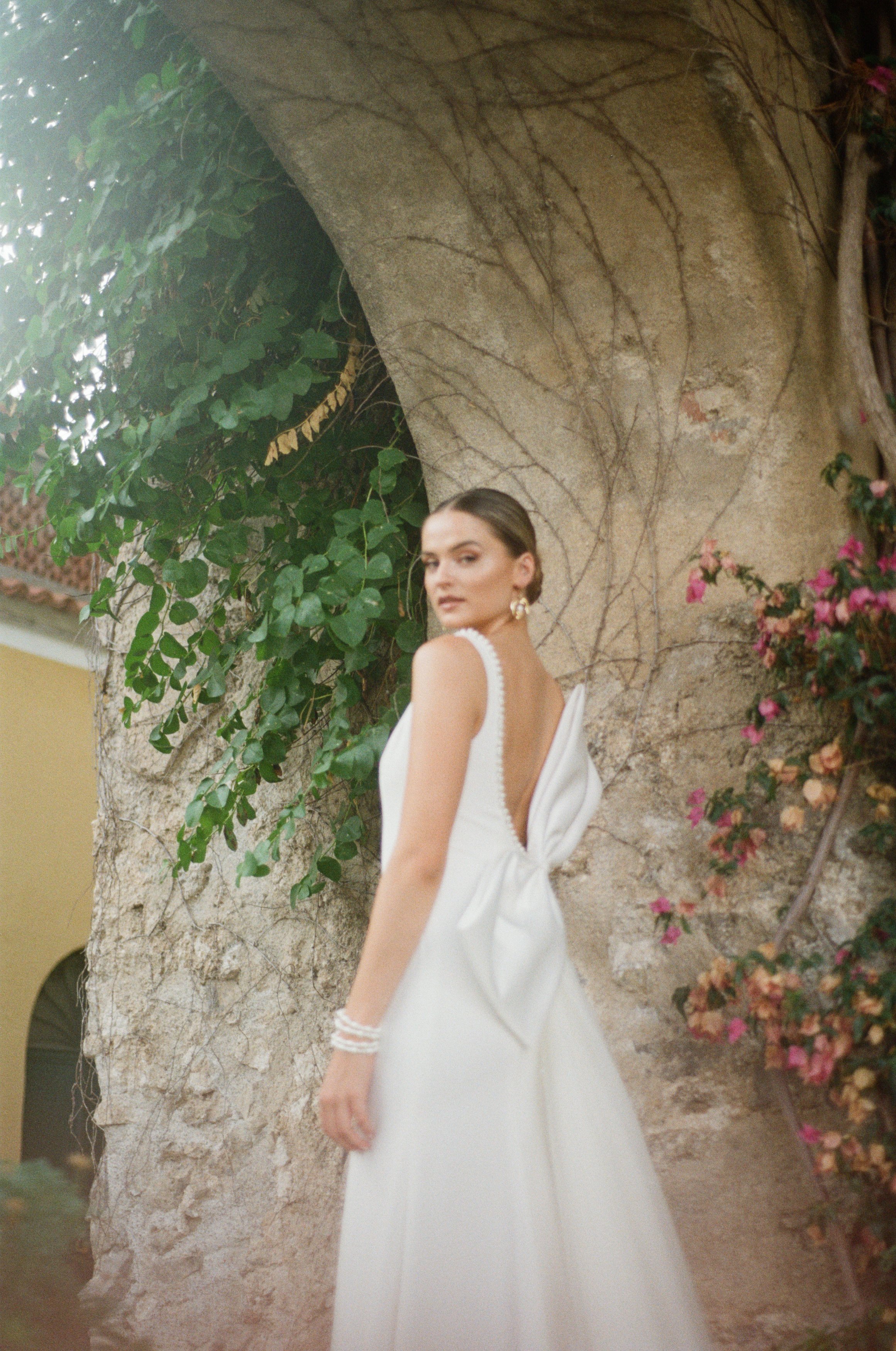 A woman in an elegant white dress with a bow back, wearing gold earrings and bracelets, standing by a tree with vines and pink flowers.