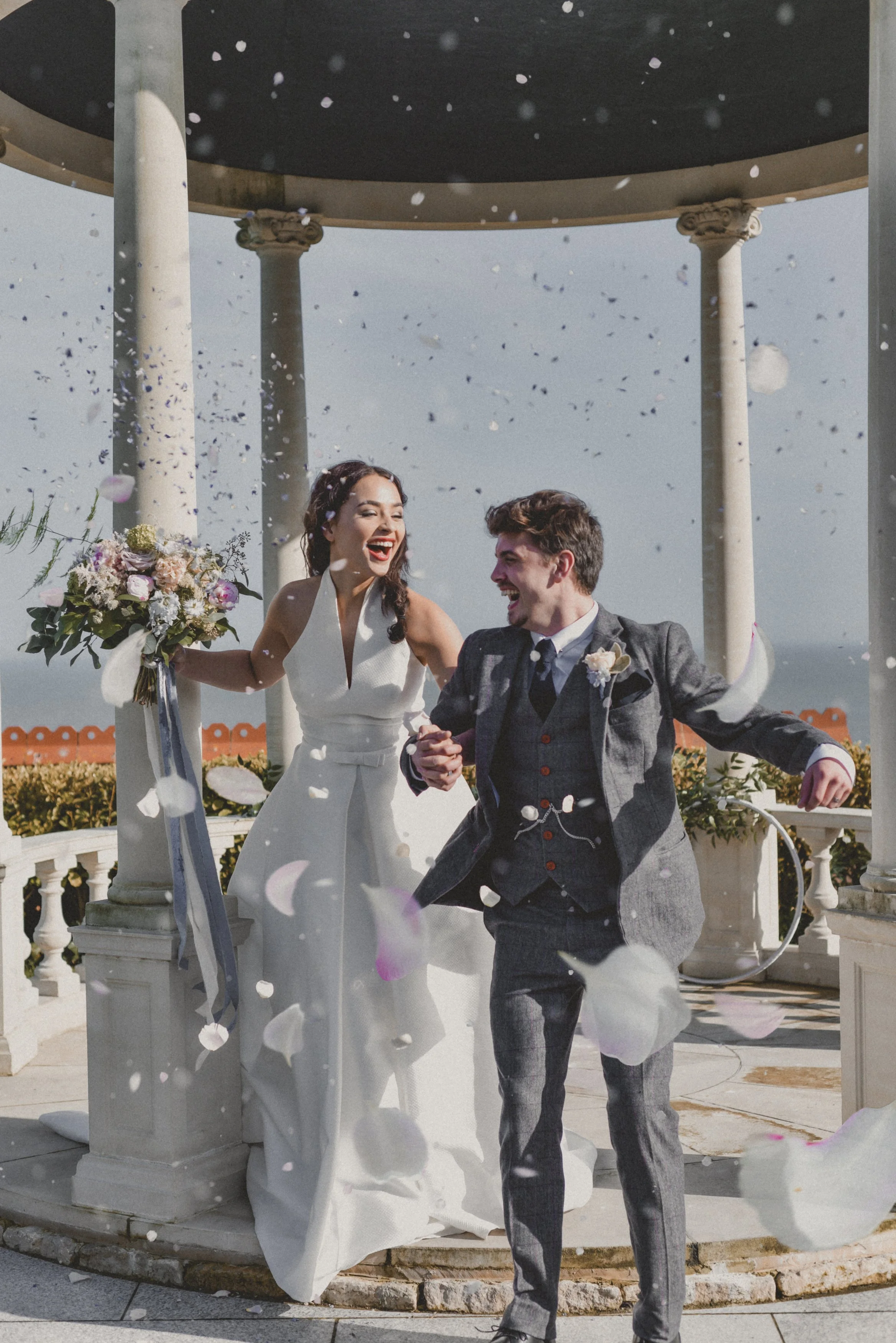 A newlywed couple celebrating their wedding outdoors under a gazebo with a view of the ocean, throwing flower petals in the air, smiling and holding hands.