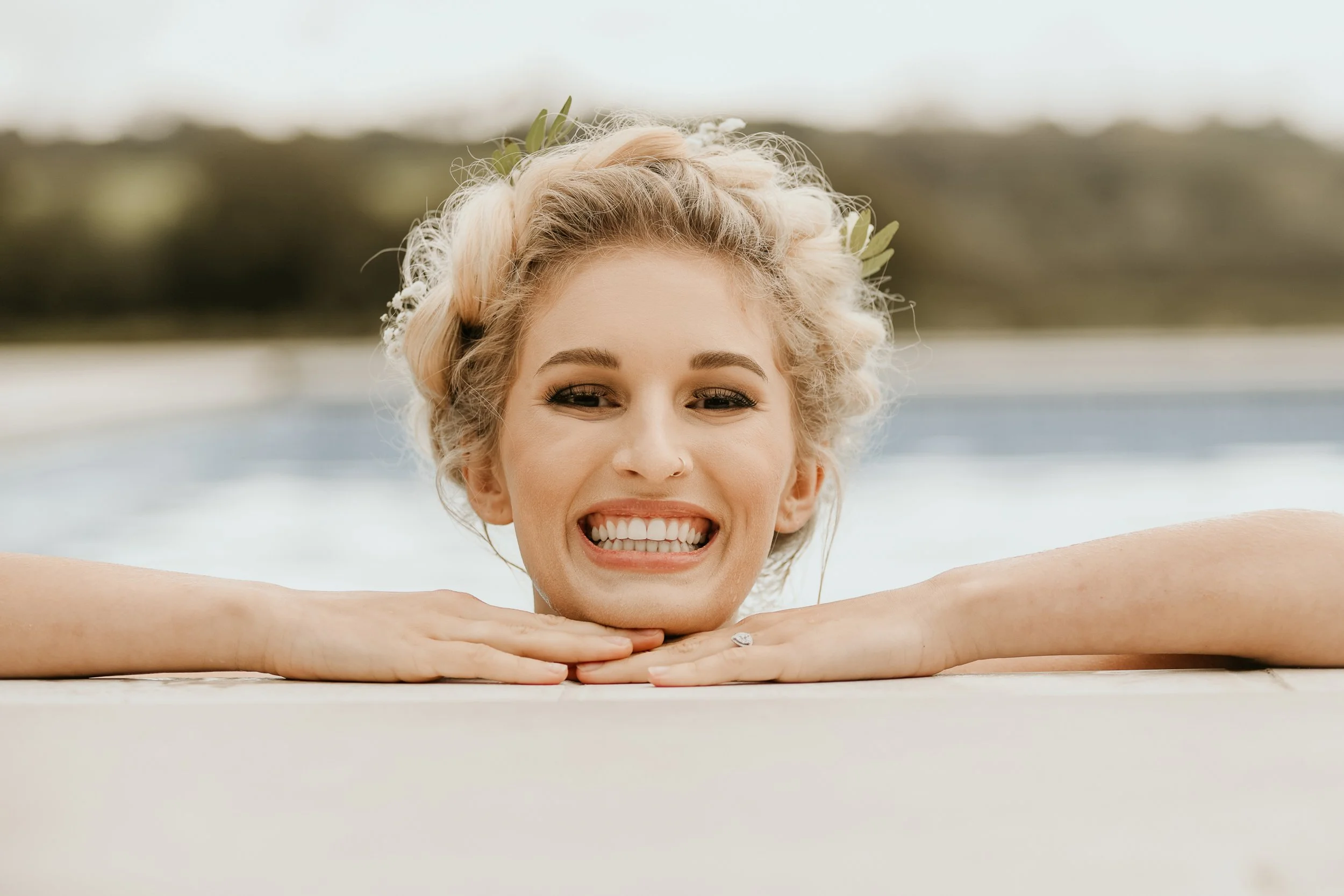 Woman with blonde, curly hair smiling with her chin resting on her hands by a pool.