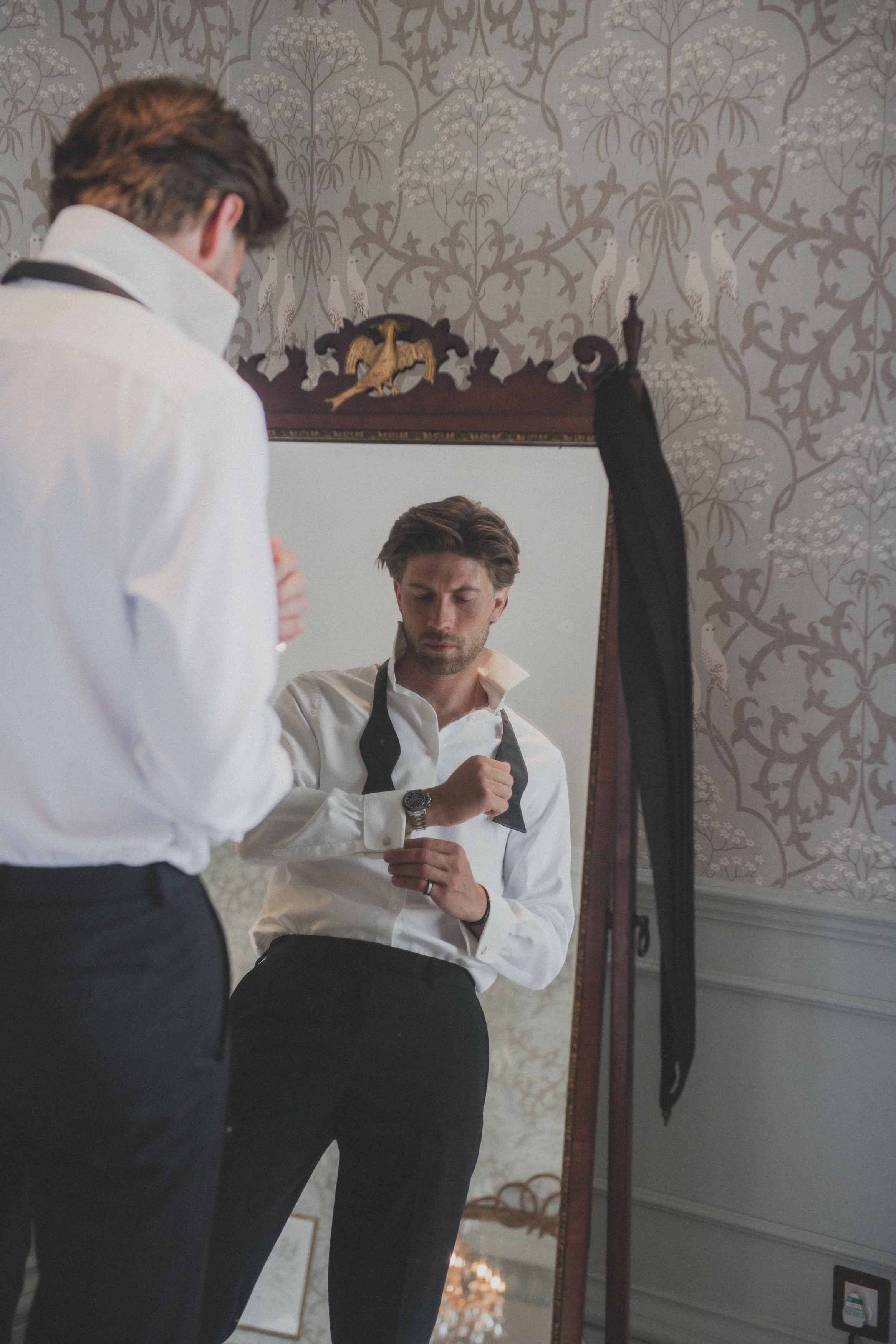 Man in white dress shirt adjusting black bow tie while looking in mirror, with wallpaper and decorative wall hanging in background.
