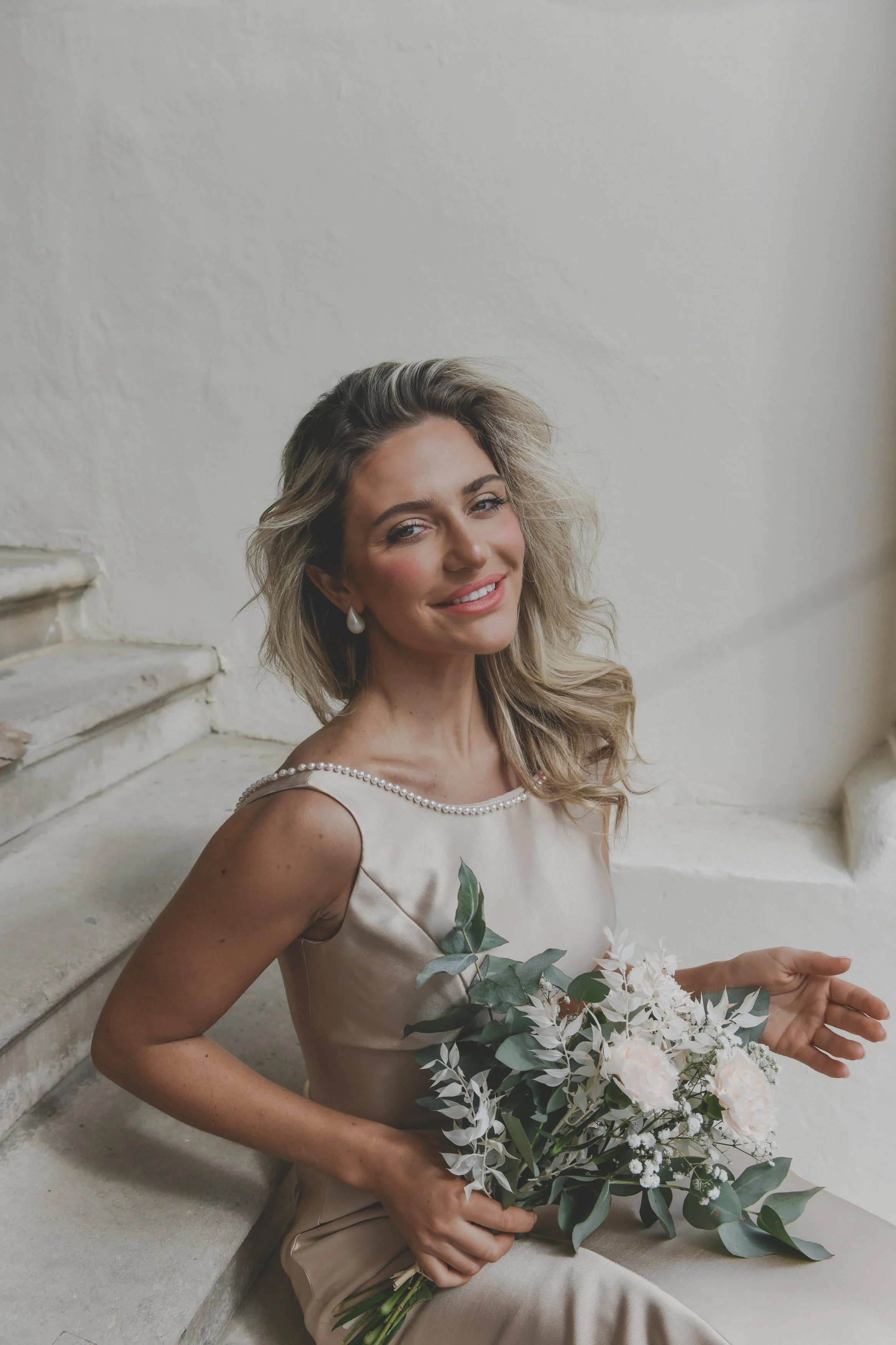 A woman with wavy blonde hair, wearing a beige dress with pearl details, holds a bouquet of white and green flowers, sitting on stairs against a white wall.