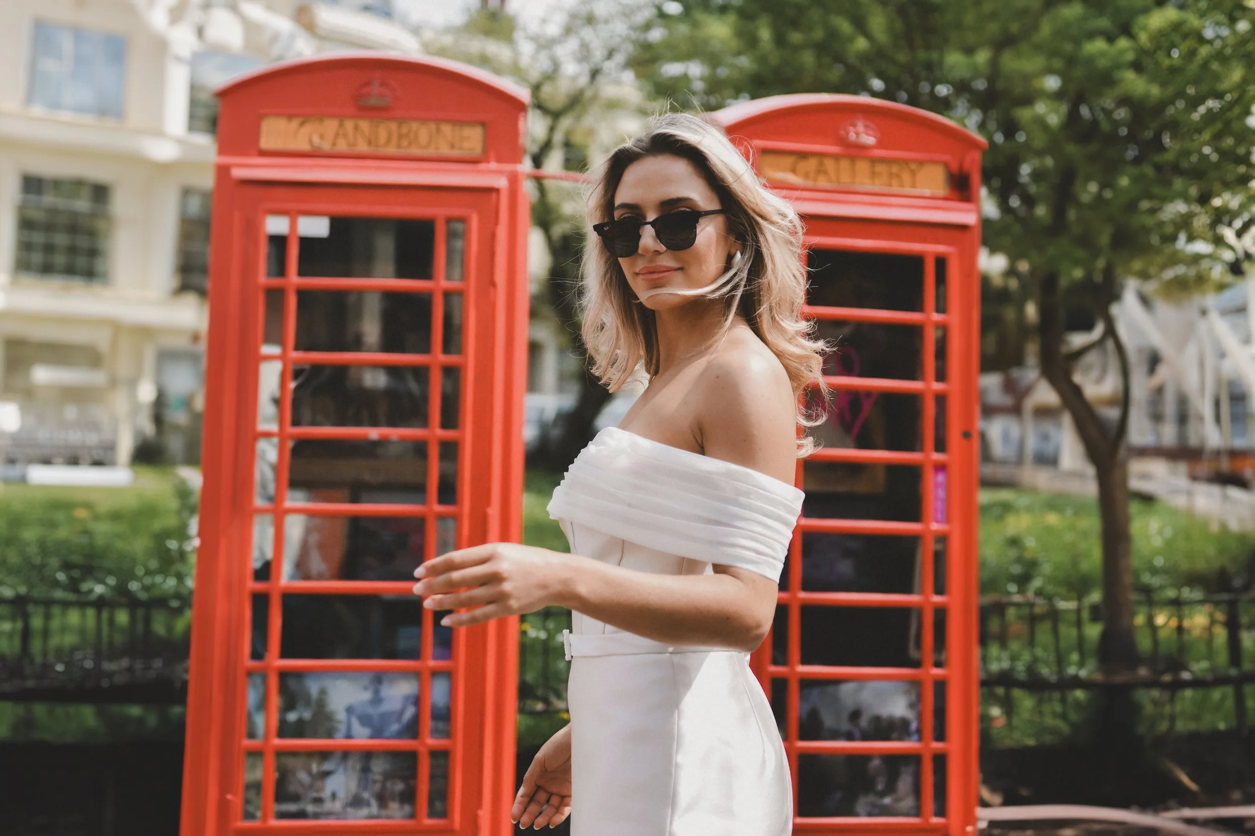 A bridesmaid in a white, off-shoulder dress and sunglasses standing in front of two red British telephone booths outdoors.