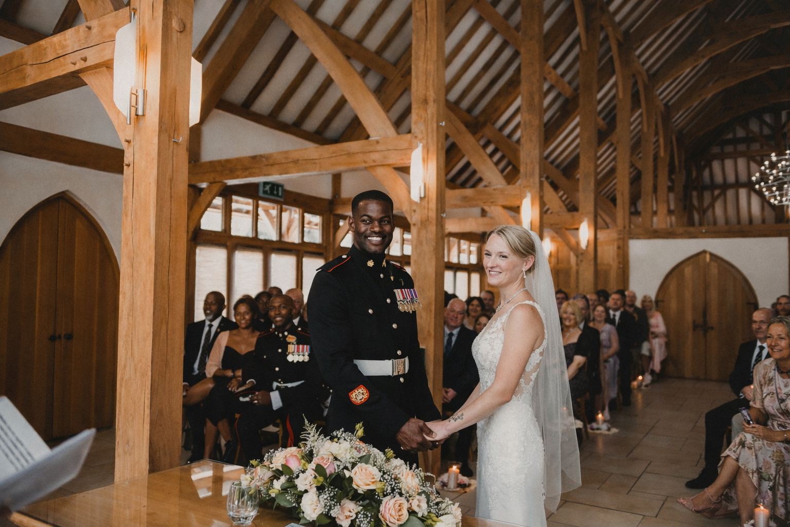 A couple getting married, with the groom in a military uniform and the bride in a white wedding dress, standing hand in hand in front of seated guests inside a wooden venue with high vaulted ceilings.
