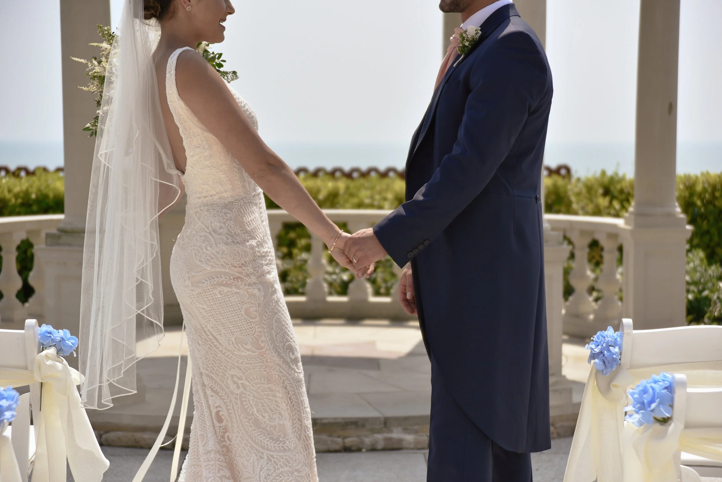 Bride and groom holding hands during their wedding ceremony outdoors, with decorated chairs and a scenic backdrop.