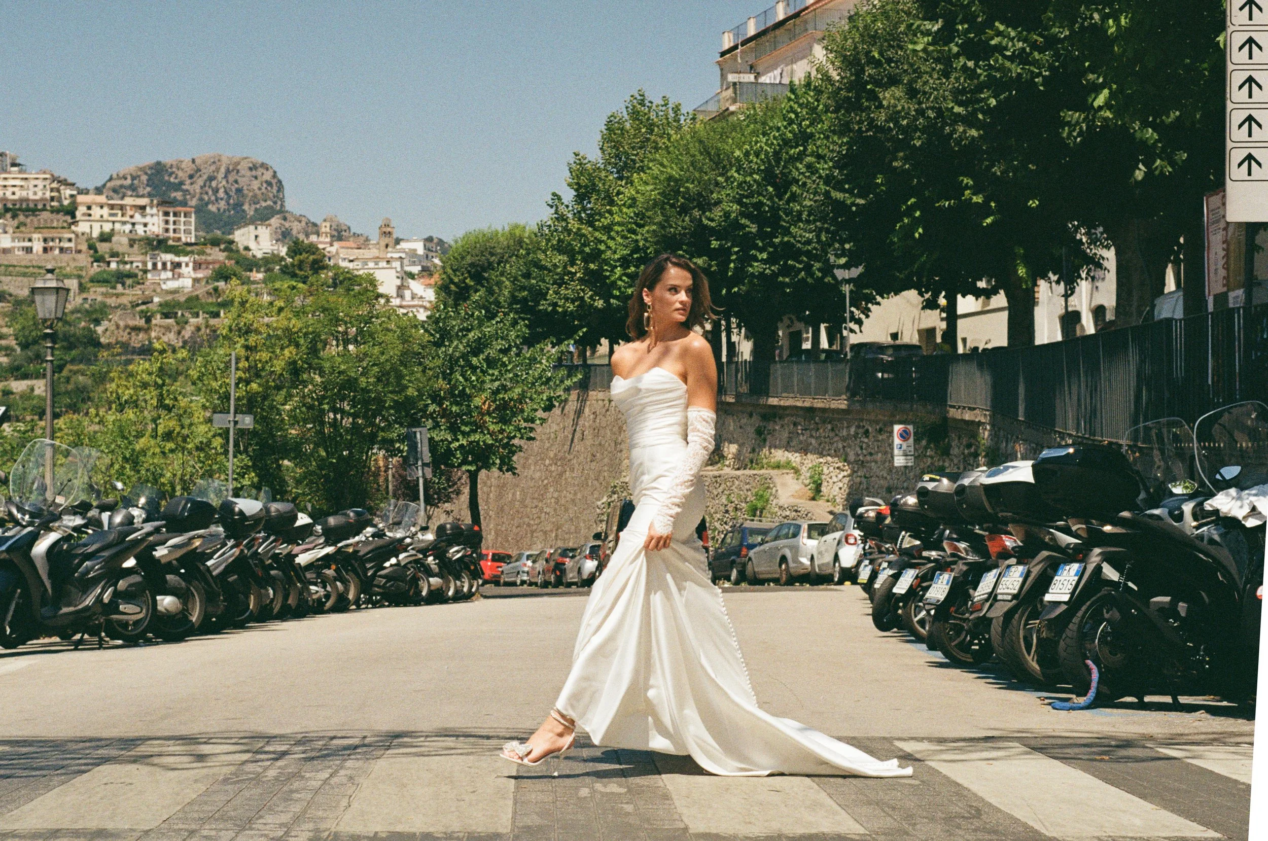 A woman in a white wedding dress and lace gloves walking across a crosswalk on a sunny day, with parked motorcycles and trees along the street.