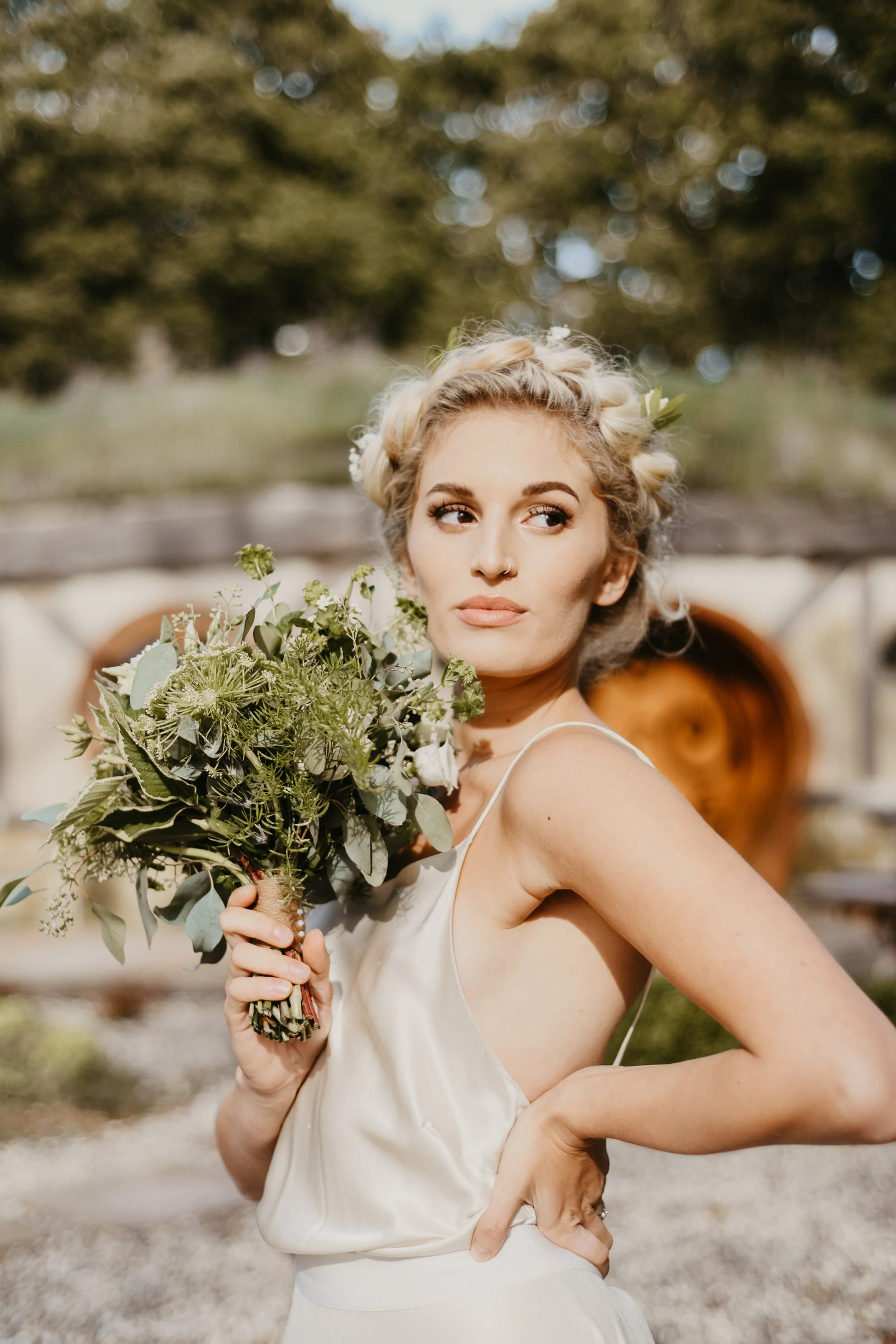 A woman with blonde, curly hair styled in an updo, holding a bouquet of green foliage and white flowers, standing outdoors with a blurred background of greenery and a wooden structure.