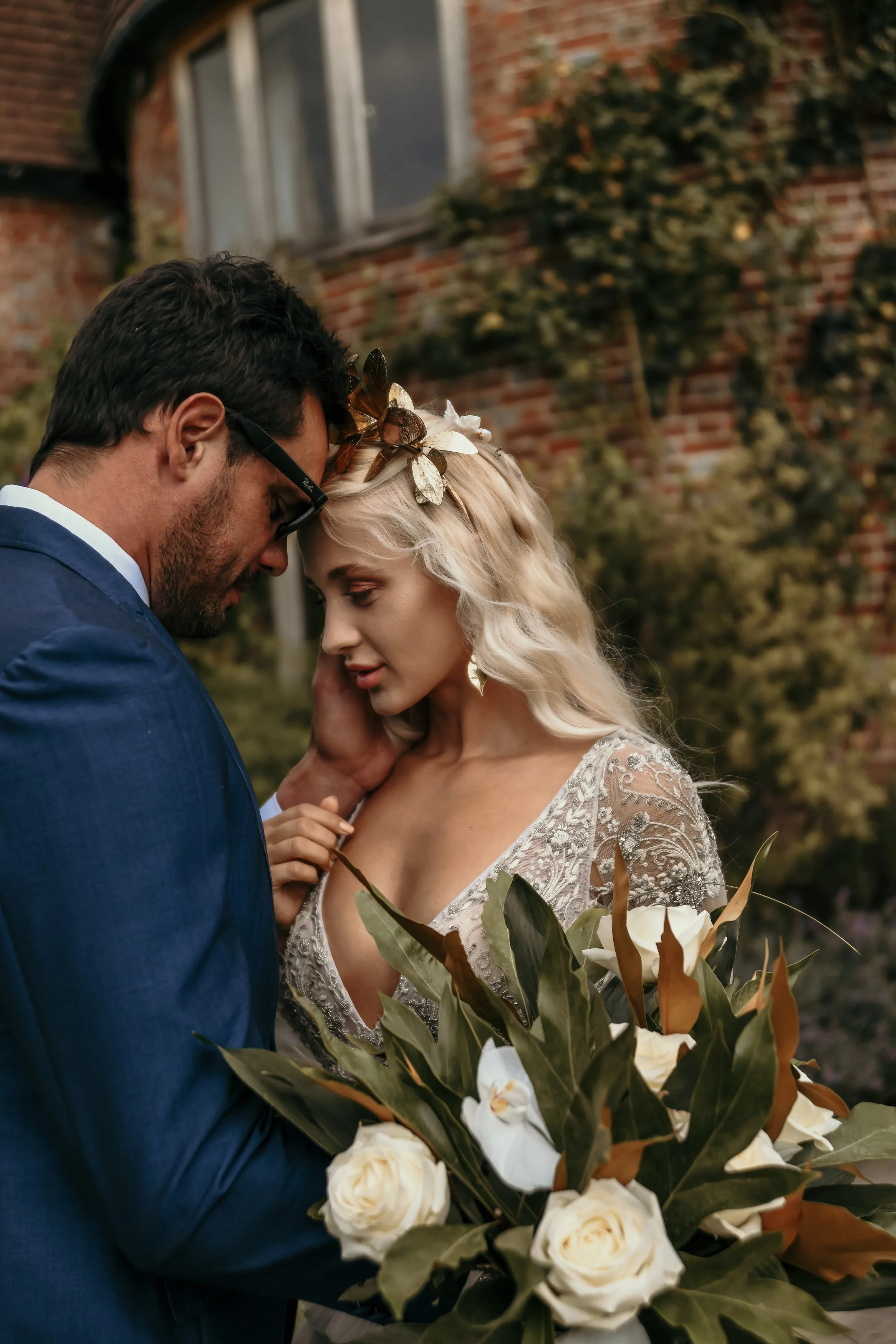 A couple dressed in wedding attire, standing close and touching foreheads, outdoors in front of a brick building with greenery, with the bride holding a large bouquet of white flowers and green leaves.