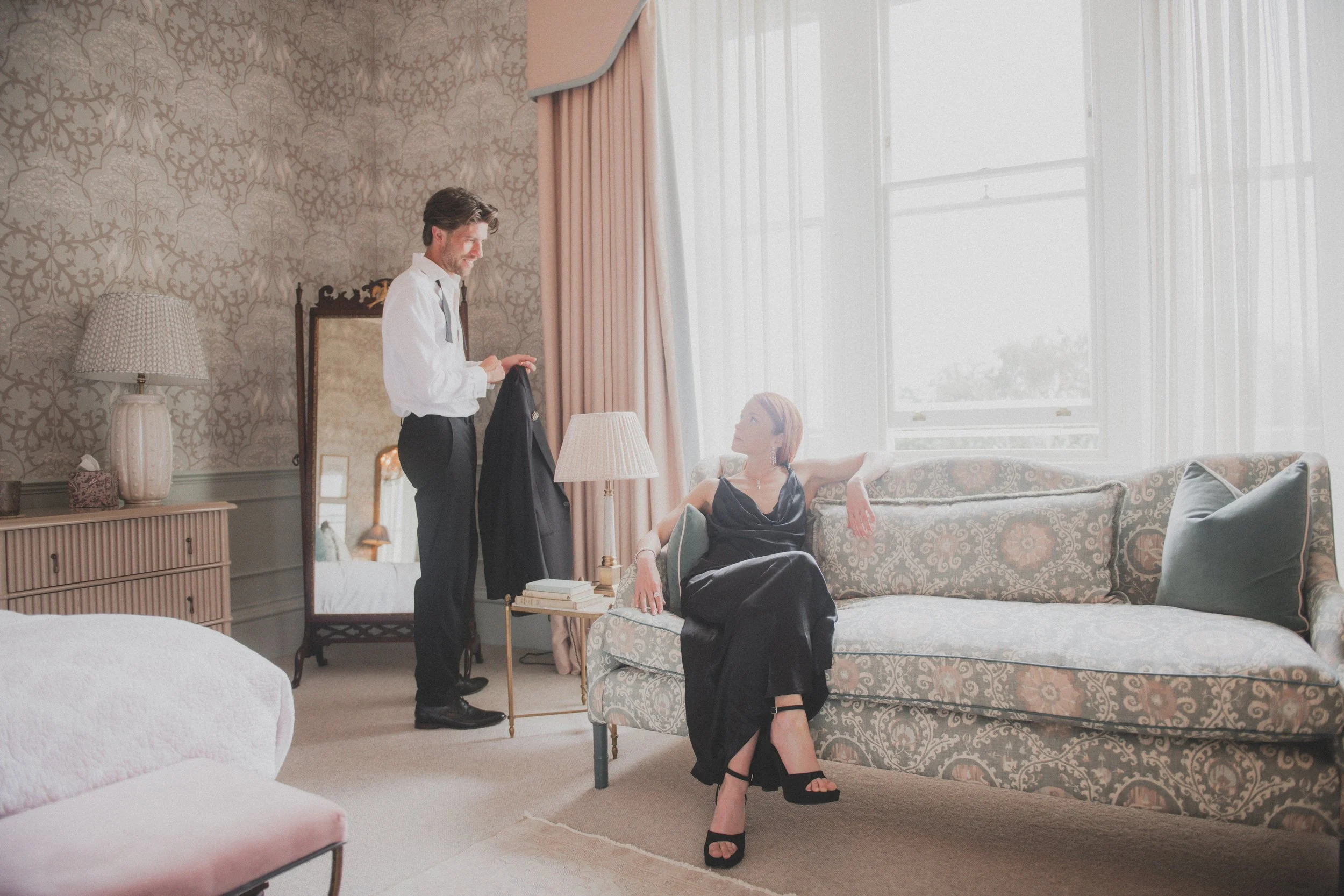 A man in a white shirt and black pants holding a black jacket stands near a woman wearing a black dress sitting on a patterned sofa, in a sunlit living room with patterned wallpaper, a mirror, side table, books, lamps, and large windows.