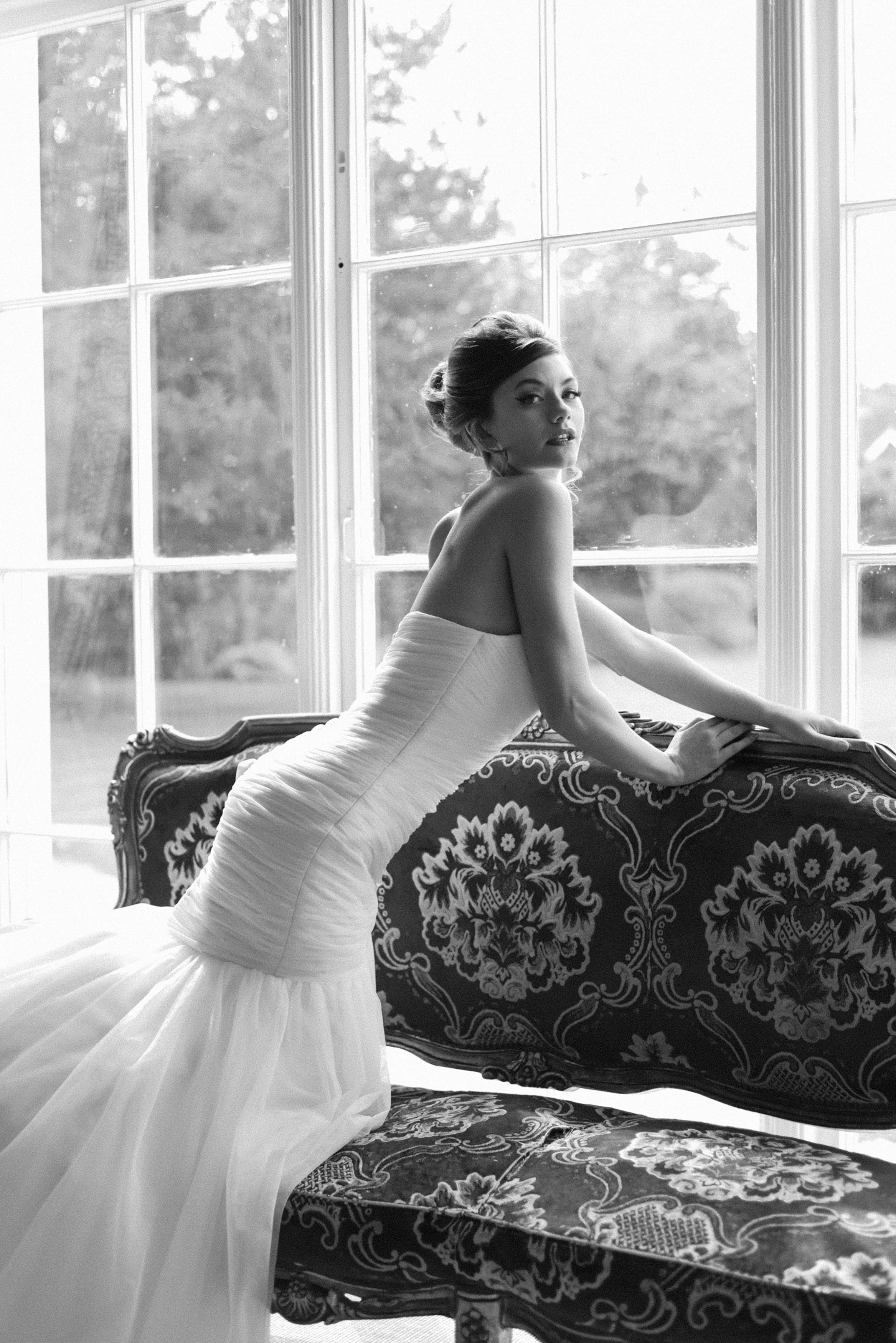 A woman in a strapless white wedding dress poses by a large window, leaning on a decorative sofa.