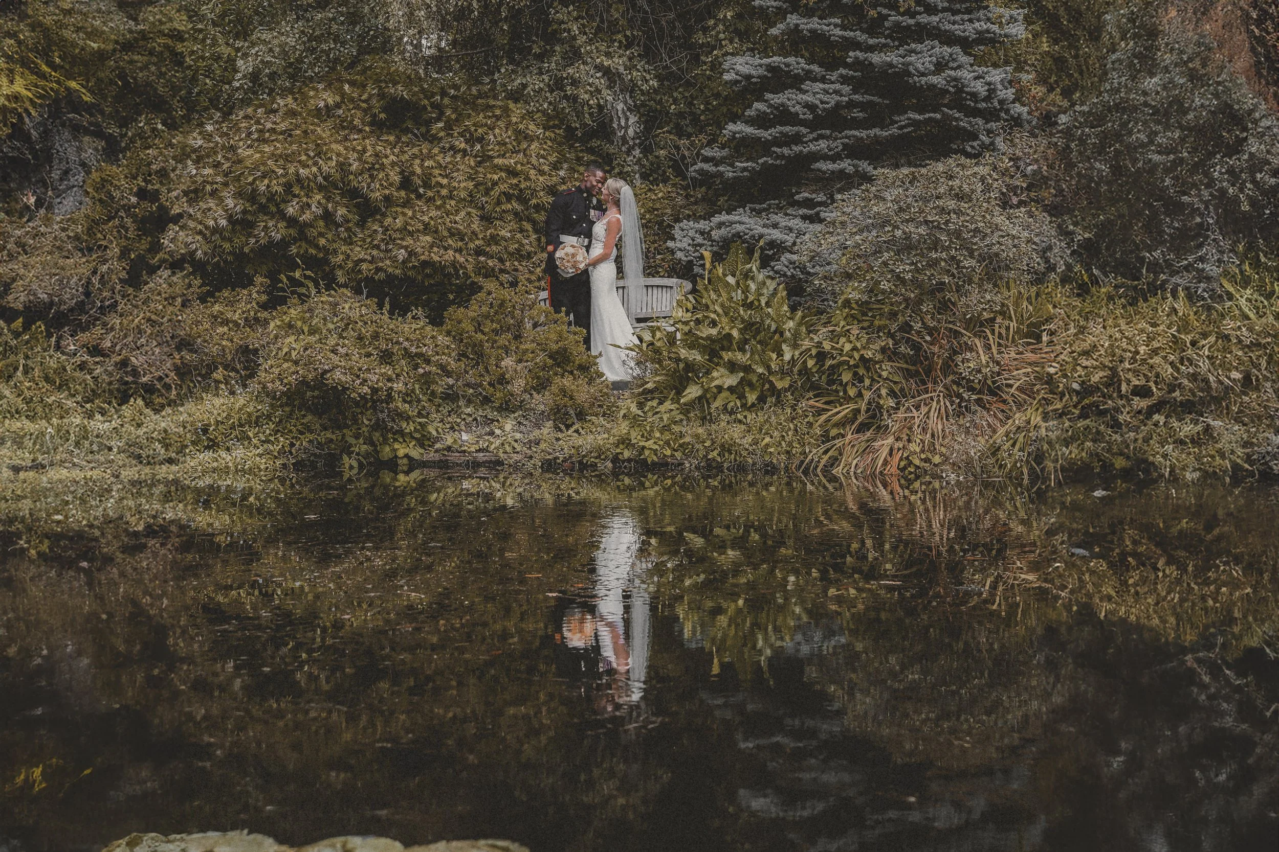 A bride and groom standing close together on a small bridge surrounded by lush greenery and trees, with their reflection visible in the water below.