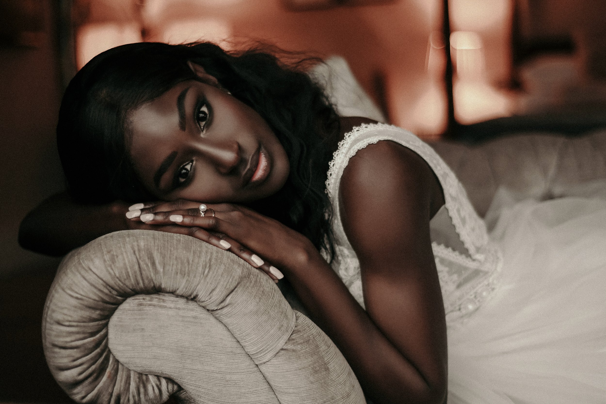 A woman with dark hair and dark skin is lying down on a beige, textured armchair with her head resting on her arm. She is wearing a white lace dress and has makeup on. The background is softly lit with warm tones.