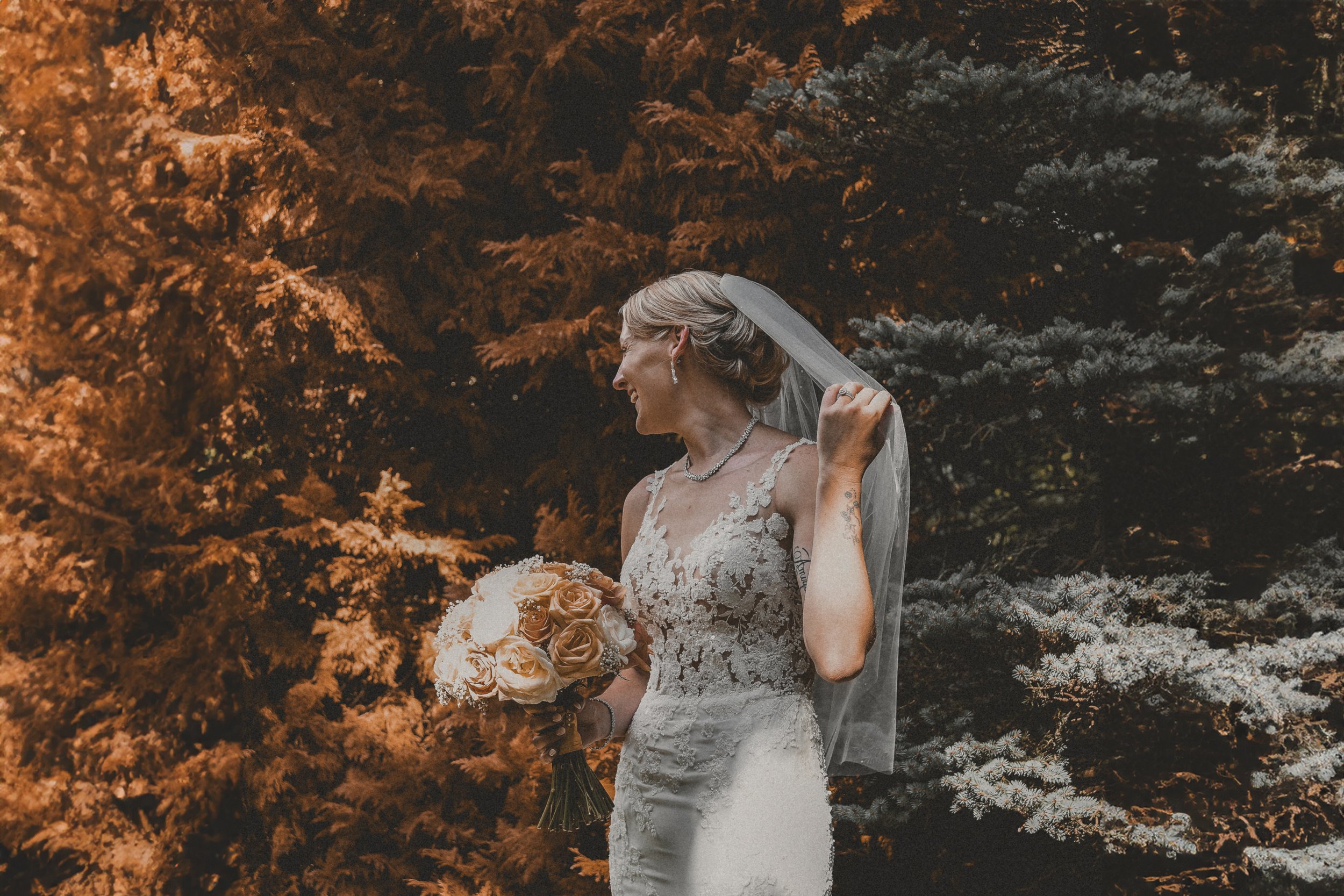 A bride in a lace wedding dress holding a bouquet of white and cream roses, smiling and adjusting her veil while standing outdoors in front of evergreen trees.