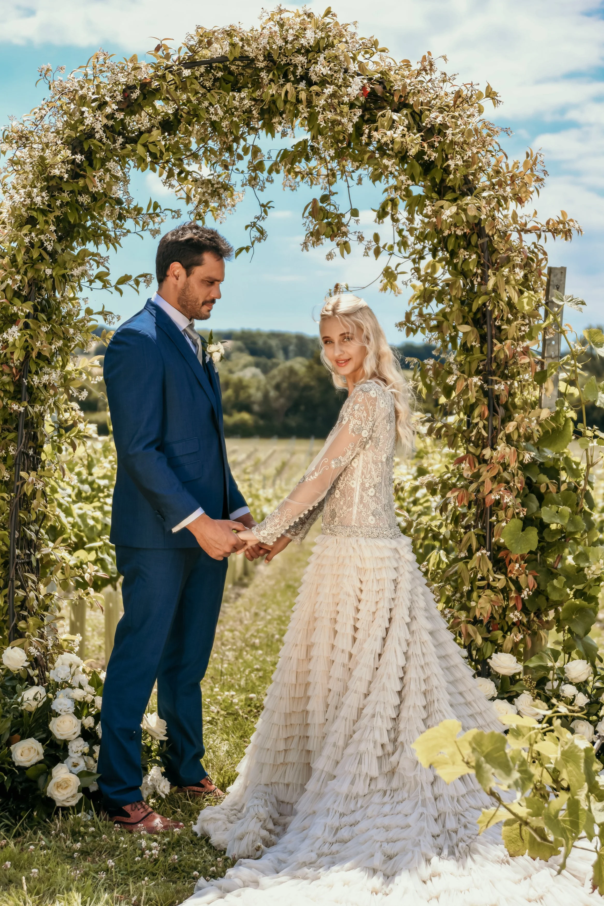 A couple holding hands and facing each other in a wedding ceremony outdoors under a floral arch, with a vineyard and blue sky in the background.