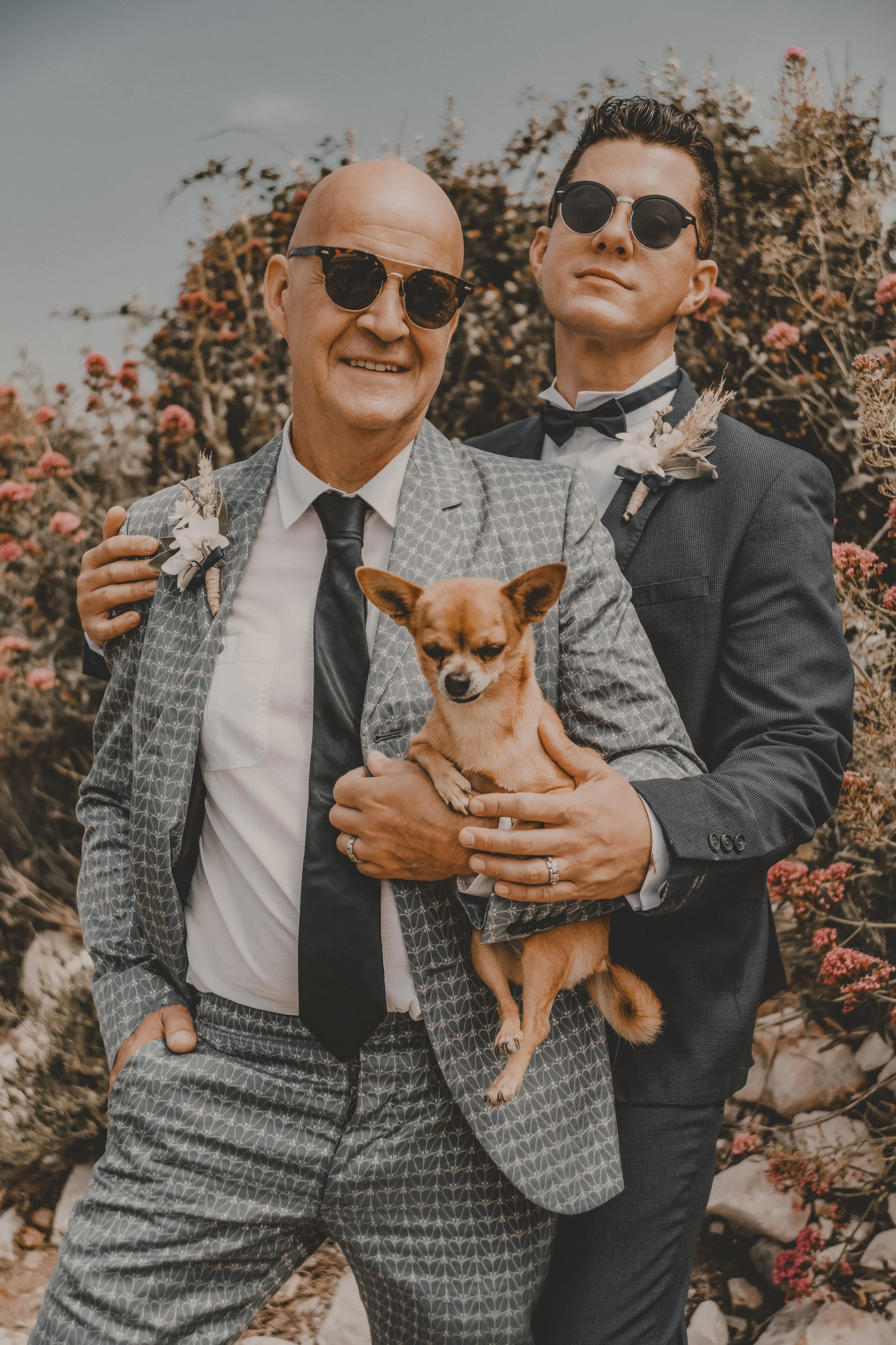 Two gay men in suits, wearing sunglasses, holding a small dog, outdoors with bushes and pink flowers in the background. Luxury gay wedding photography.