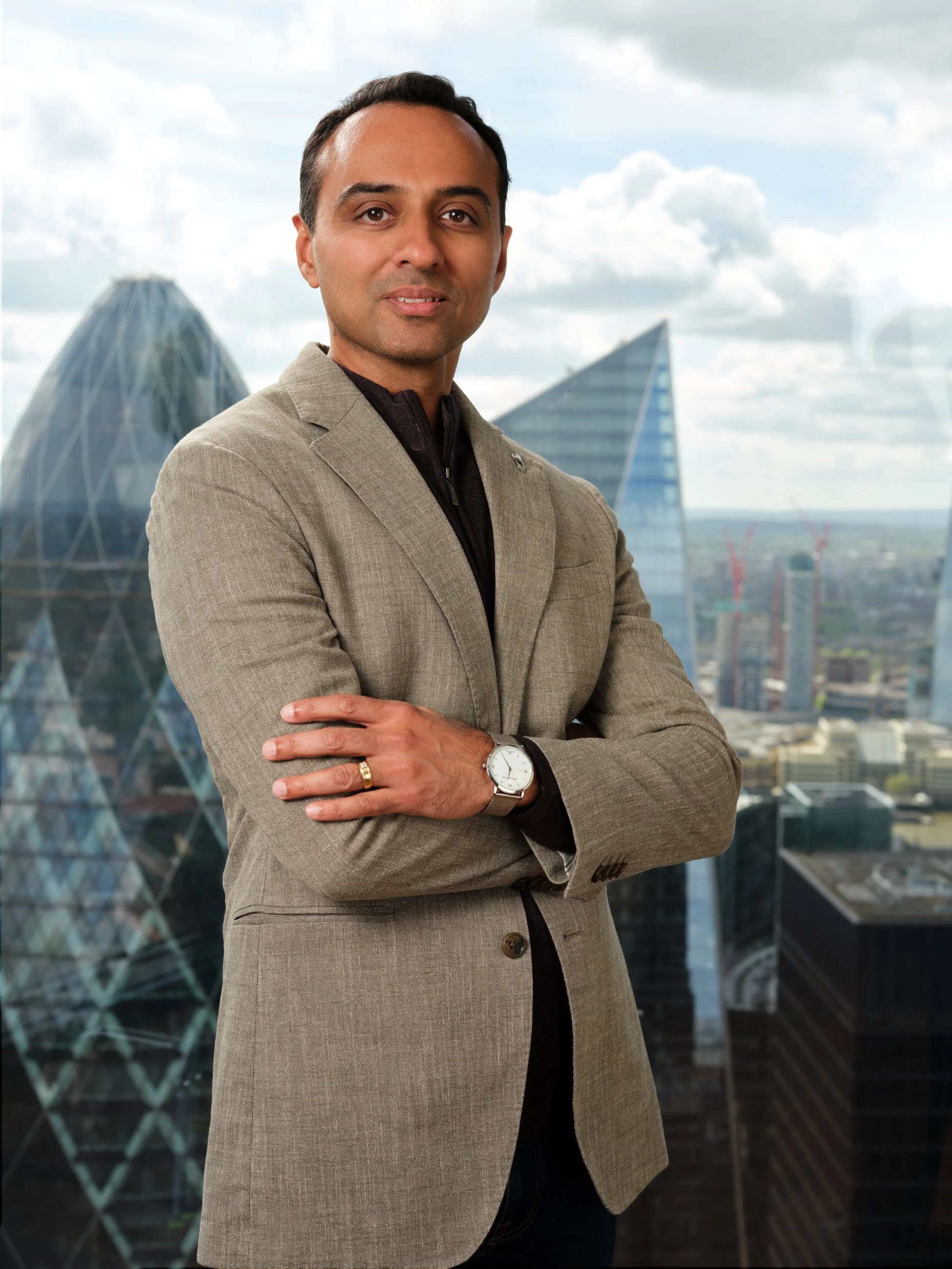 A man in a beige blazer with arms crossed, standing in front of a cityscape with modern skyscrapers.
