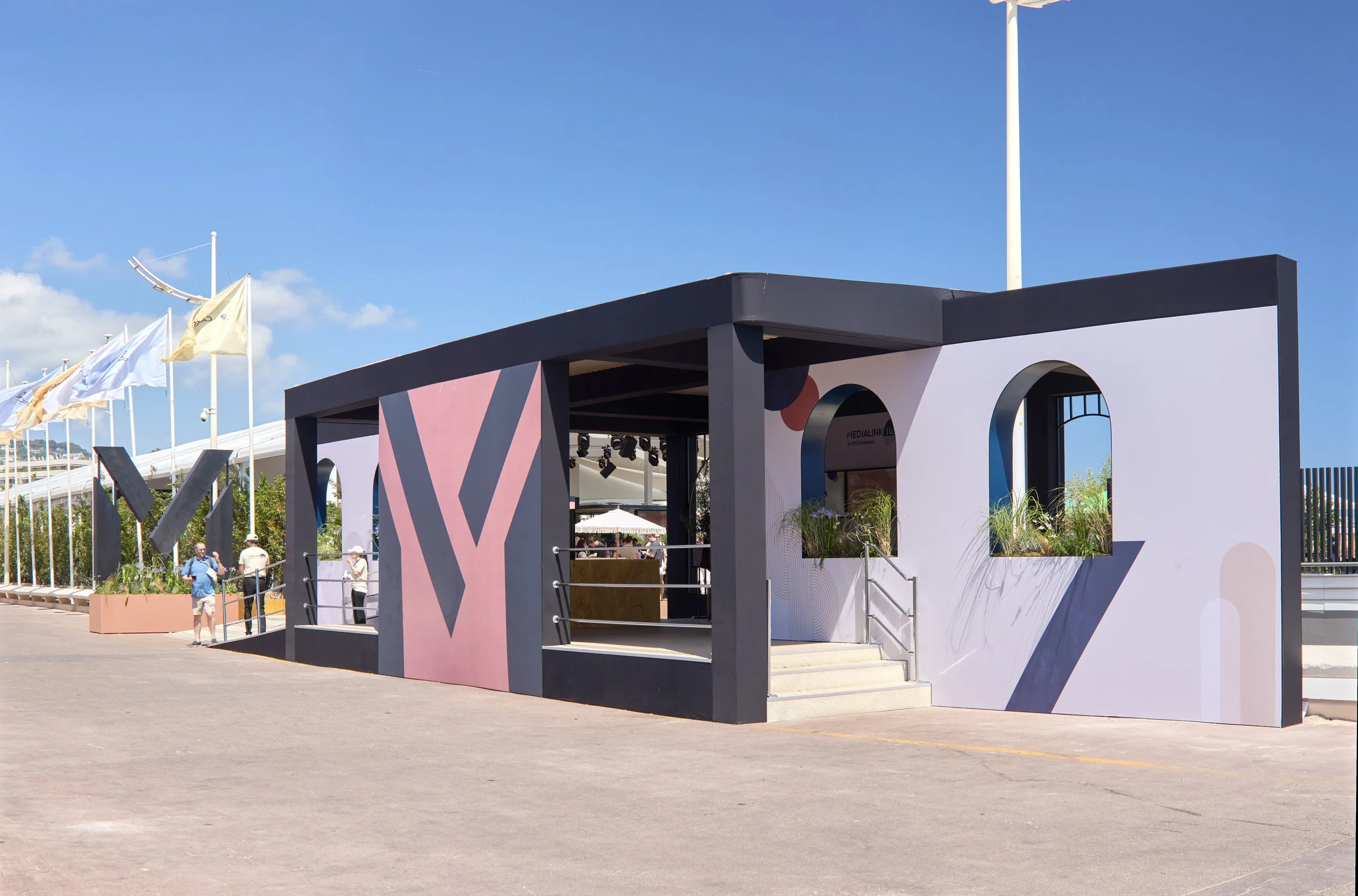 A modern booth with black and pink panels, open on one side, with stairs leading up to the entrance, and decorative plants in rectangular planters, set outdoors on a sunny day with flags flying in the background.