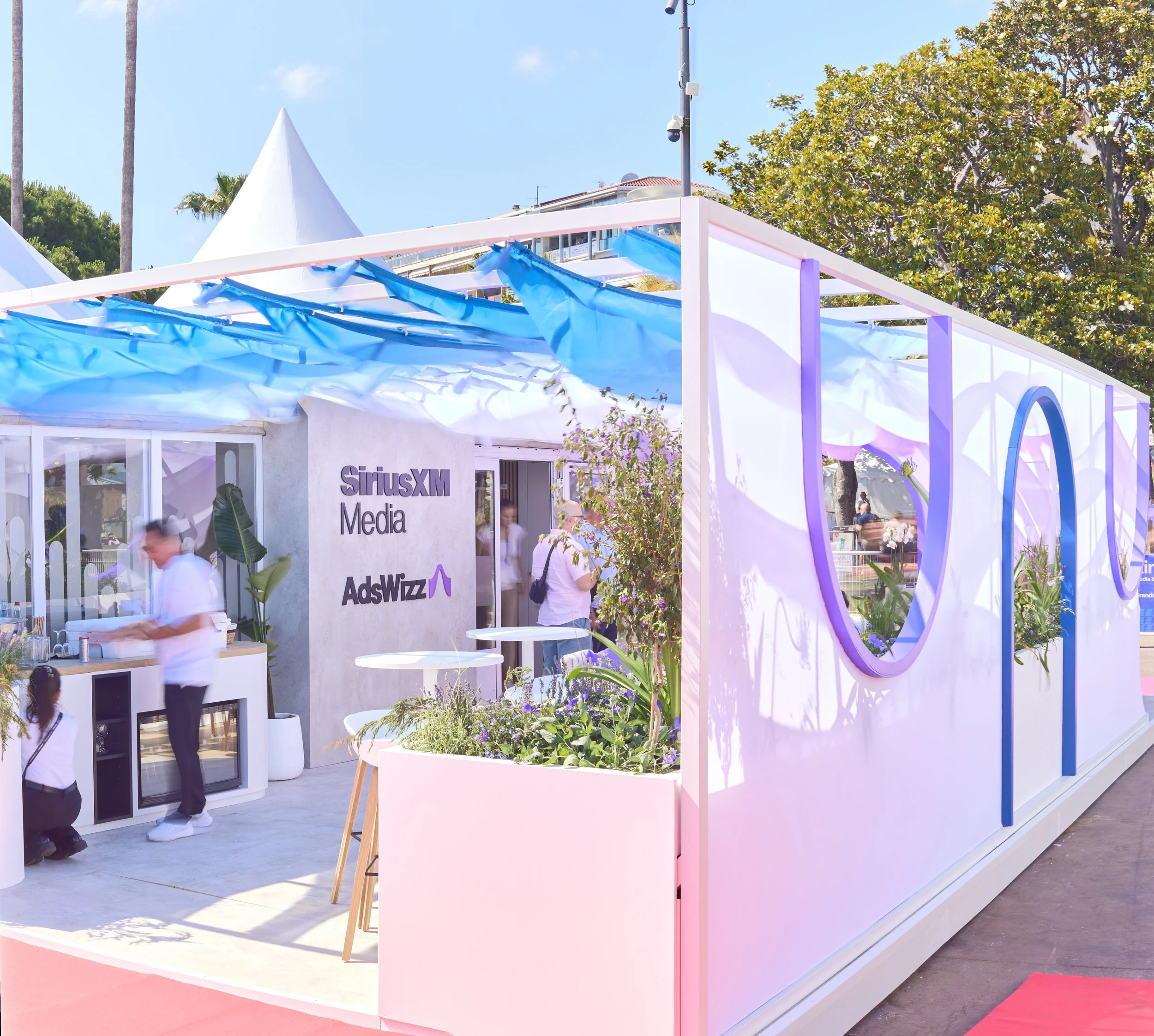 A modern outdoor event space with white and pastel-colored decor, plants, and people gathered around, featuring a sign that says 'SiriusXM Media' and 'AdsWizz'.