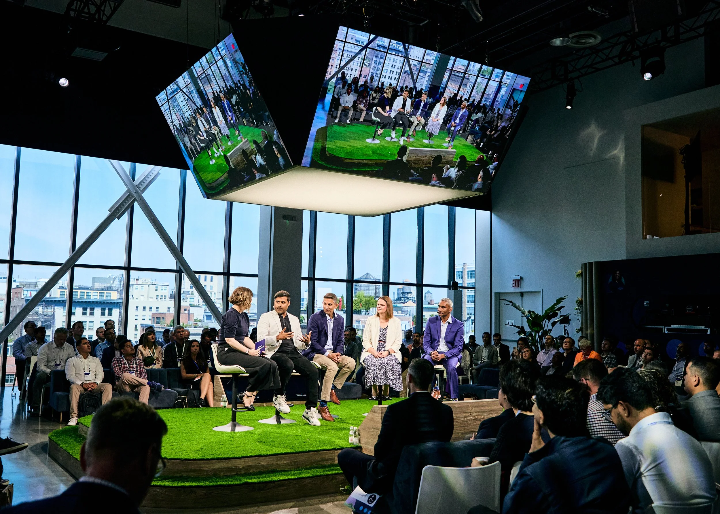 A panel discussion taking place in a modern, glass-walled conference room with a city skyline view. Five panelists, including three men and two women, are seated on a small elevated stage with a green turf carpet. A woman is speaking with a notebook,
