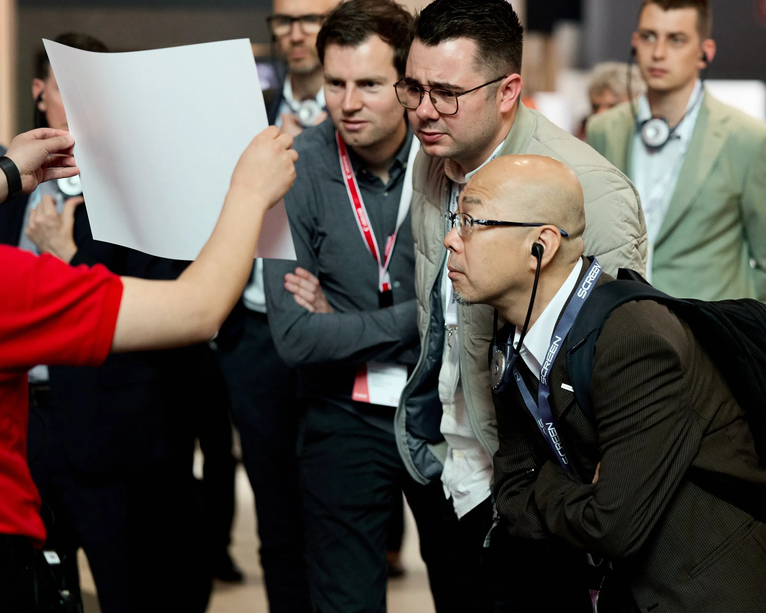Group of people at a conference or event, closely examining a blank piece of paper or document held by someone in a red shirt, with one man wearing headphones and a lanyard, and others appearing focused and engaged.