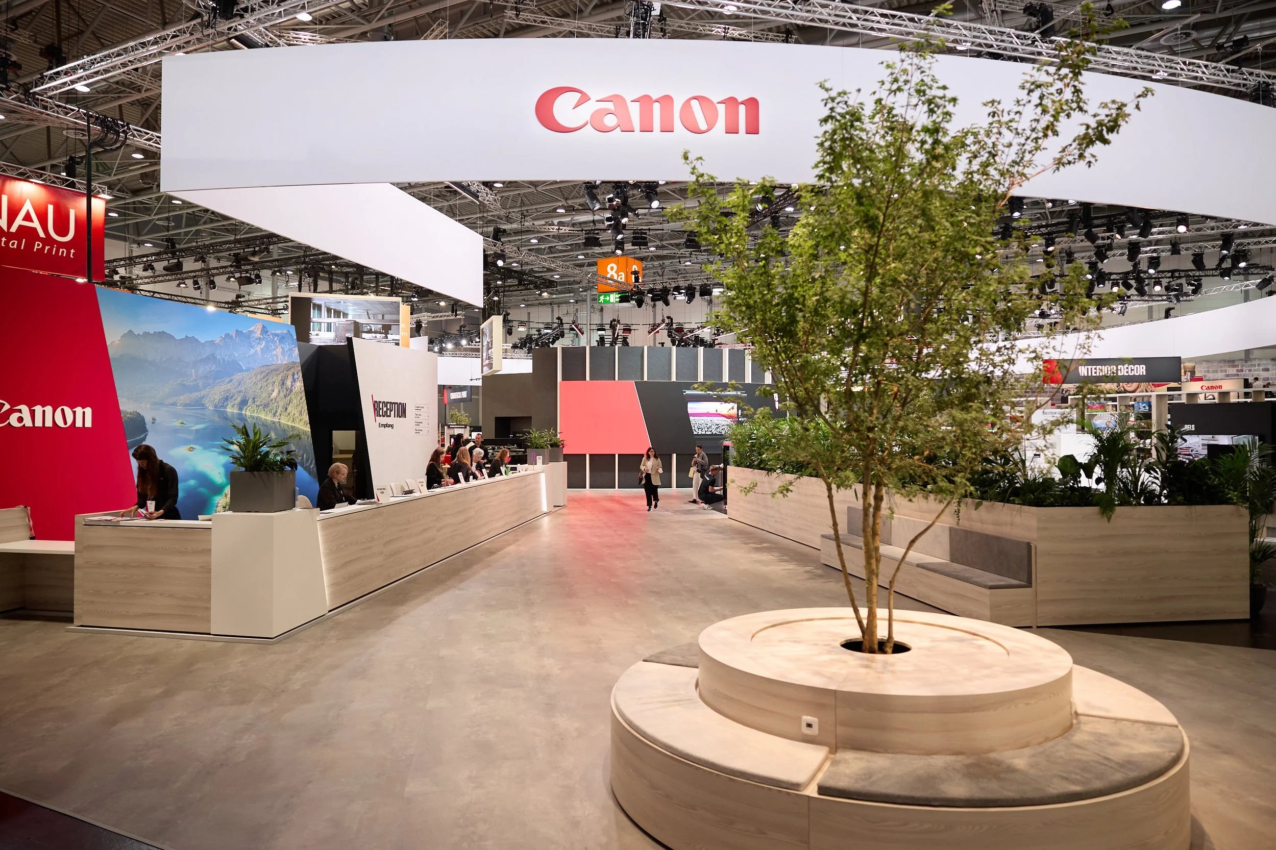 Interior of a large exhibition hall with a Canon booth, reception desk, people working and walking around, and a seating area with a tree and plants in a circular wooden planter.
