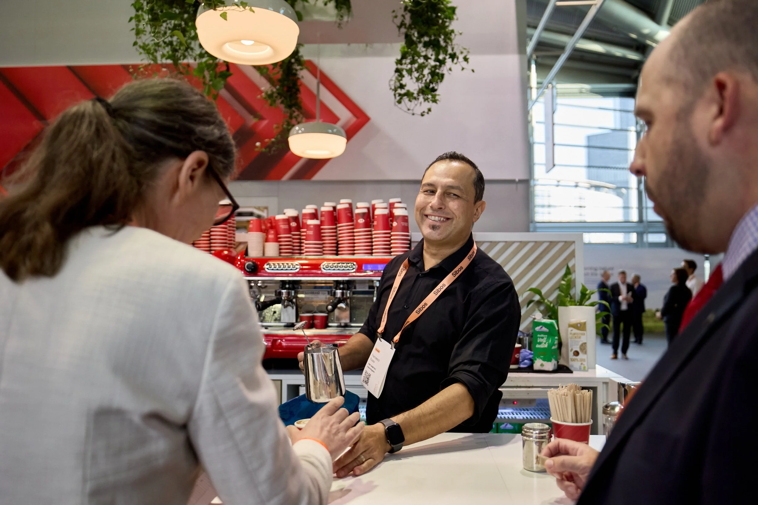 A barista smiling while serving two customers at a coffee bar in a modern, brightly lit cafe. The setting features red cups stacked behind him, a professional espresso machine, and decorative elements like plants and a geometric red wall art piece.