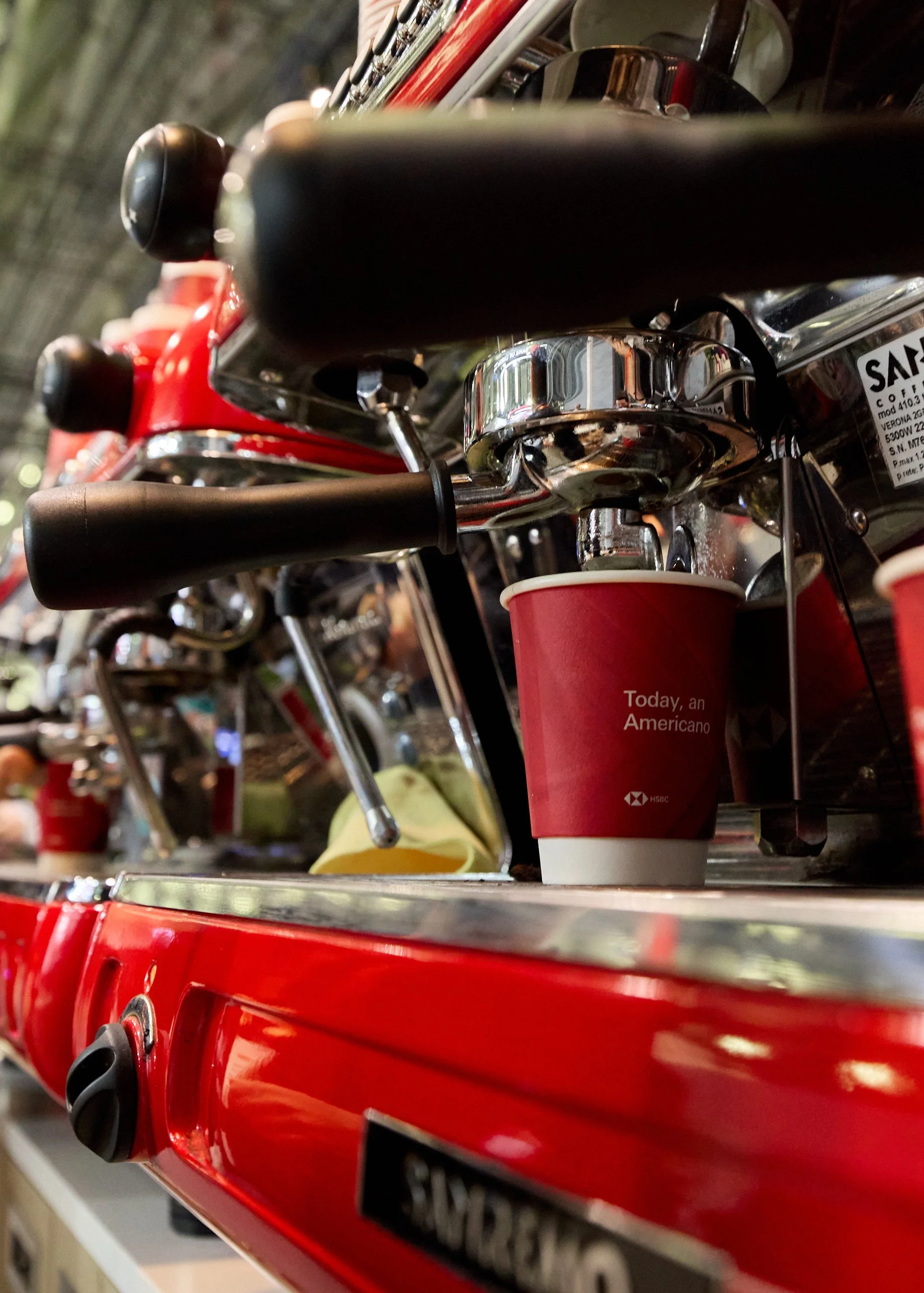 Close-up view of a red espresso machine with a portafilter attached, a red coffee cup with the text 'Today, an Americano,' and a background of other coffee machines and accessories.