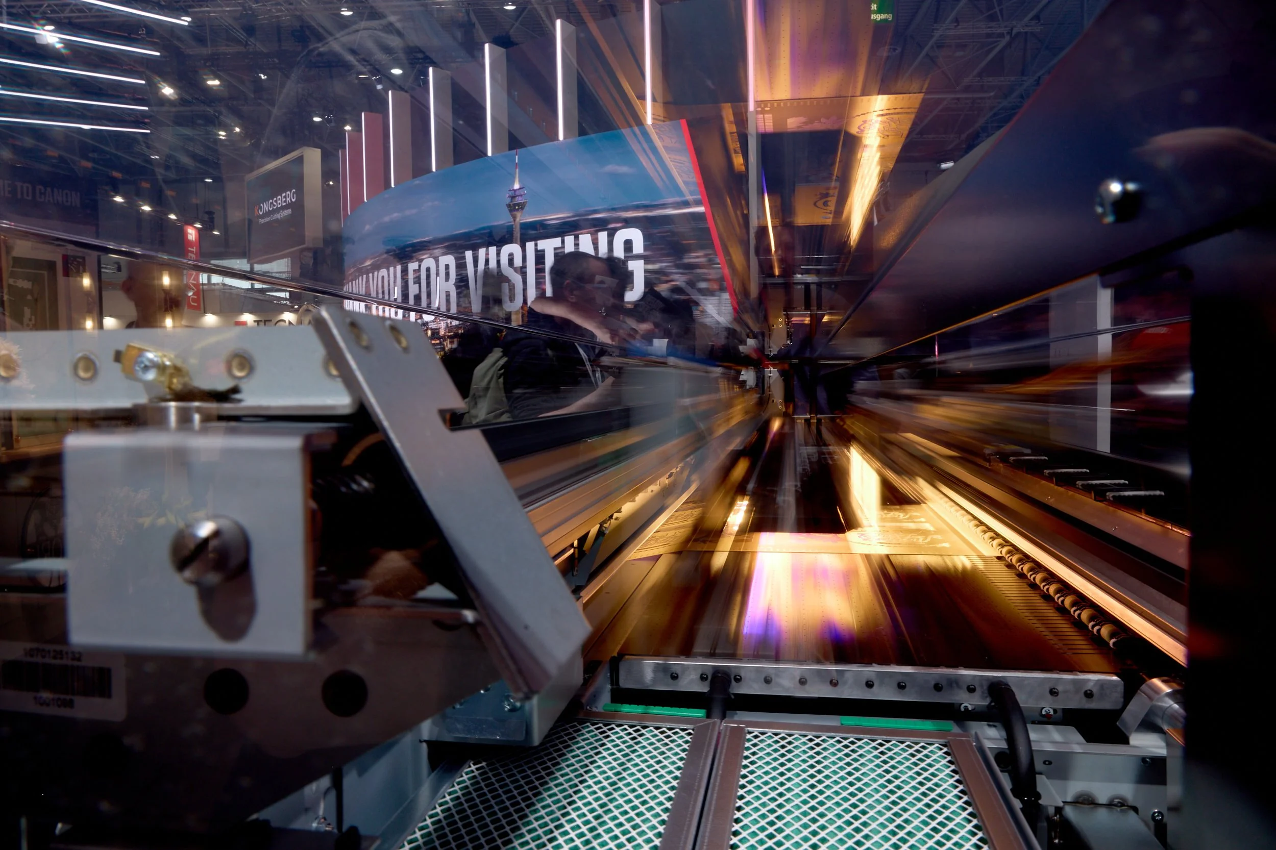 Close-up of machinery with mechanical parts, reflective surfaces, and digital display reading 'YOU' for visiting, among blurred background of people and exhibit at a technology or electronics trade show.