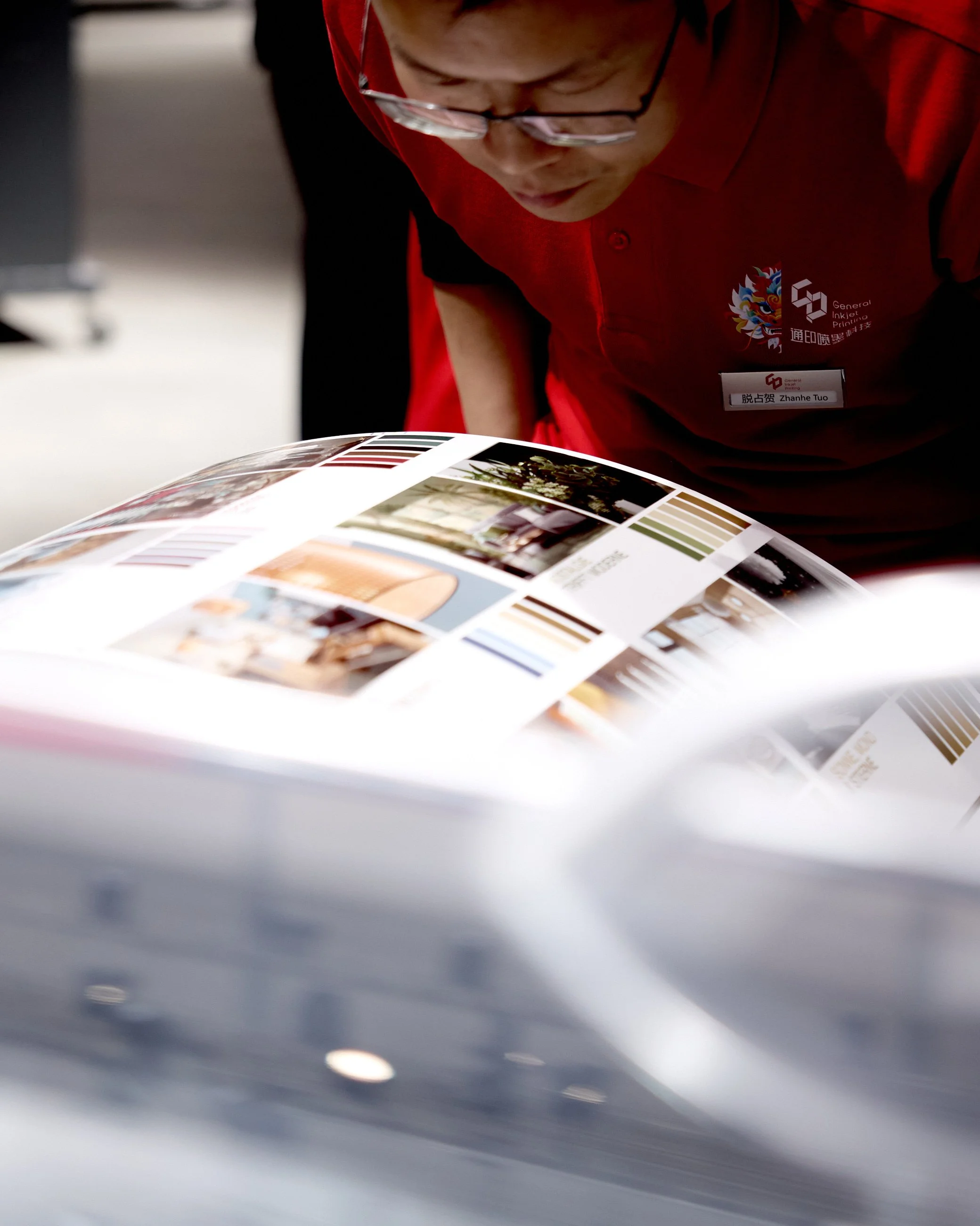 A person wearing glasses and a red shirt leaning over a large sheet of printed material with images and color swatches, likely at a printing or design fair.