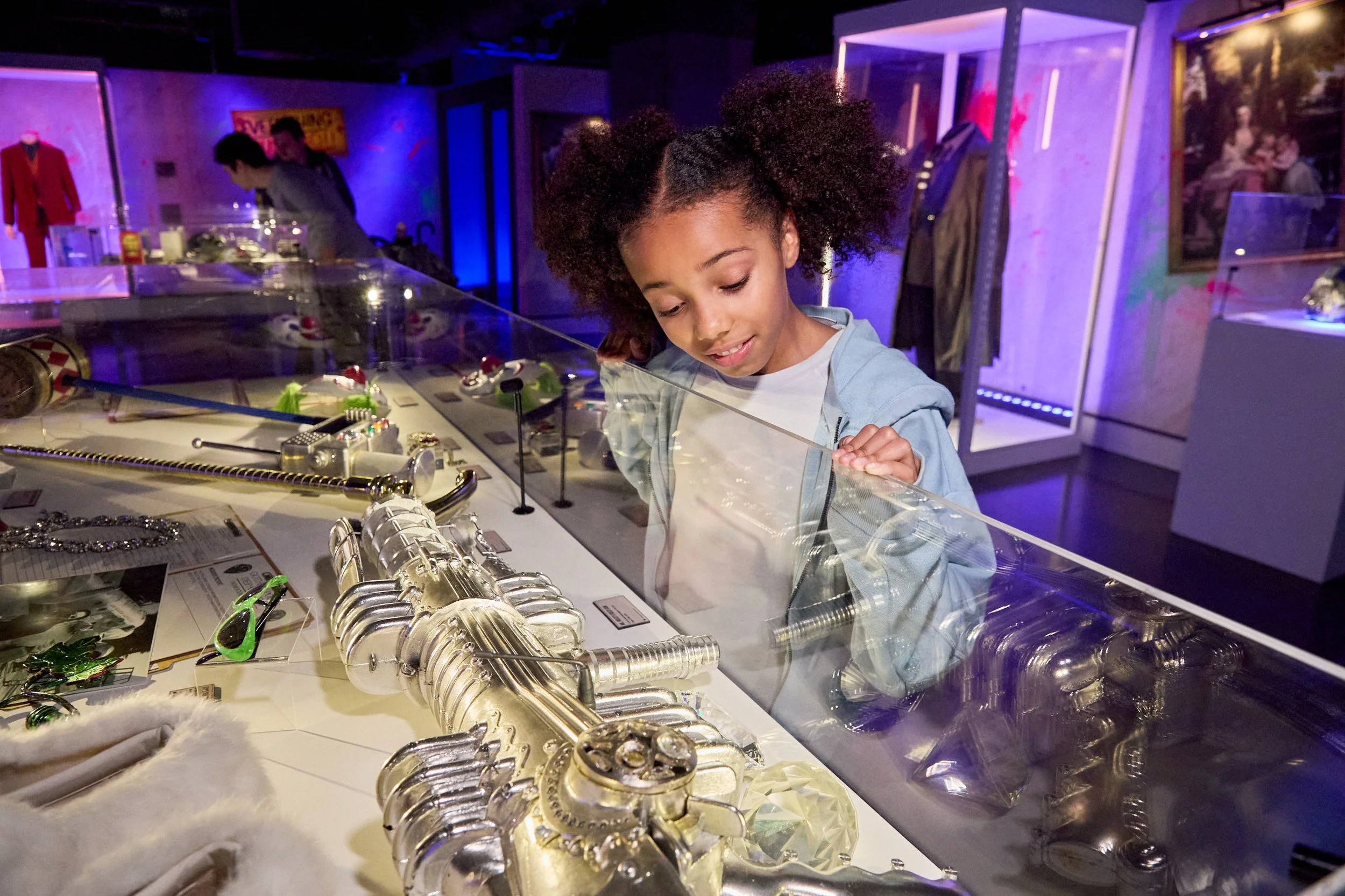A young girl with curly hair looking at jewelry and accessories displayed behind a glass case in a museum or exhibit space with purple and blue lighting.