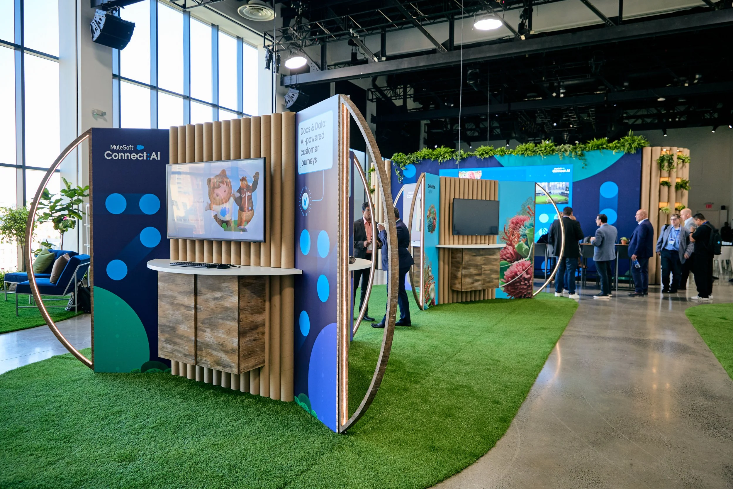Trade show booth with blue and green design, screens displaying animated characters, and a group of people in business attire at an indoor conference.