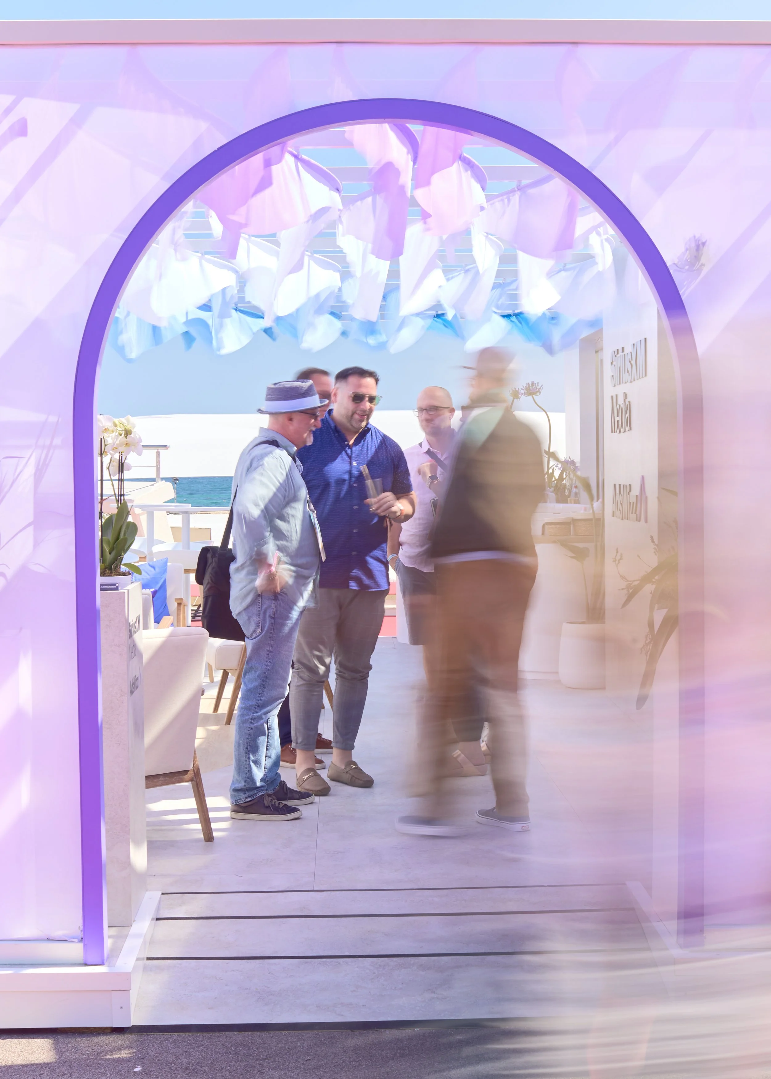 A group of four men standing and talking on a patio with an ocean view, seen through a purple-framed archway decorated with white fabric. One man wears a hat, sunglasses, and a backpack. All are casually dressed in summer clothing.
