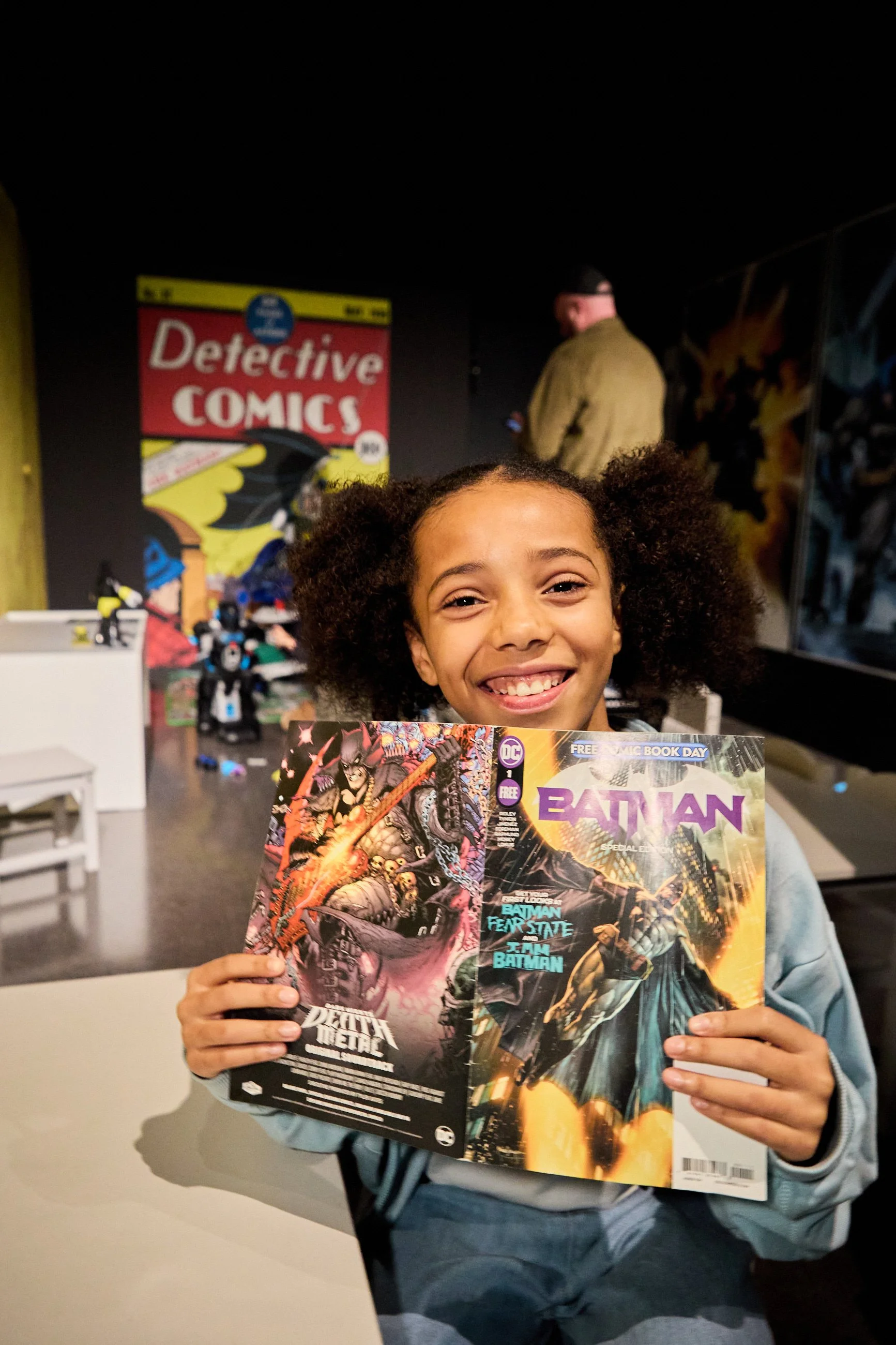 A young girl with curly hair smiling and holding up two comic books, one featuring Batman, in a comic store or convention setting.