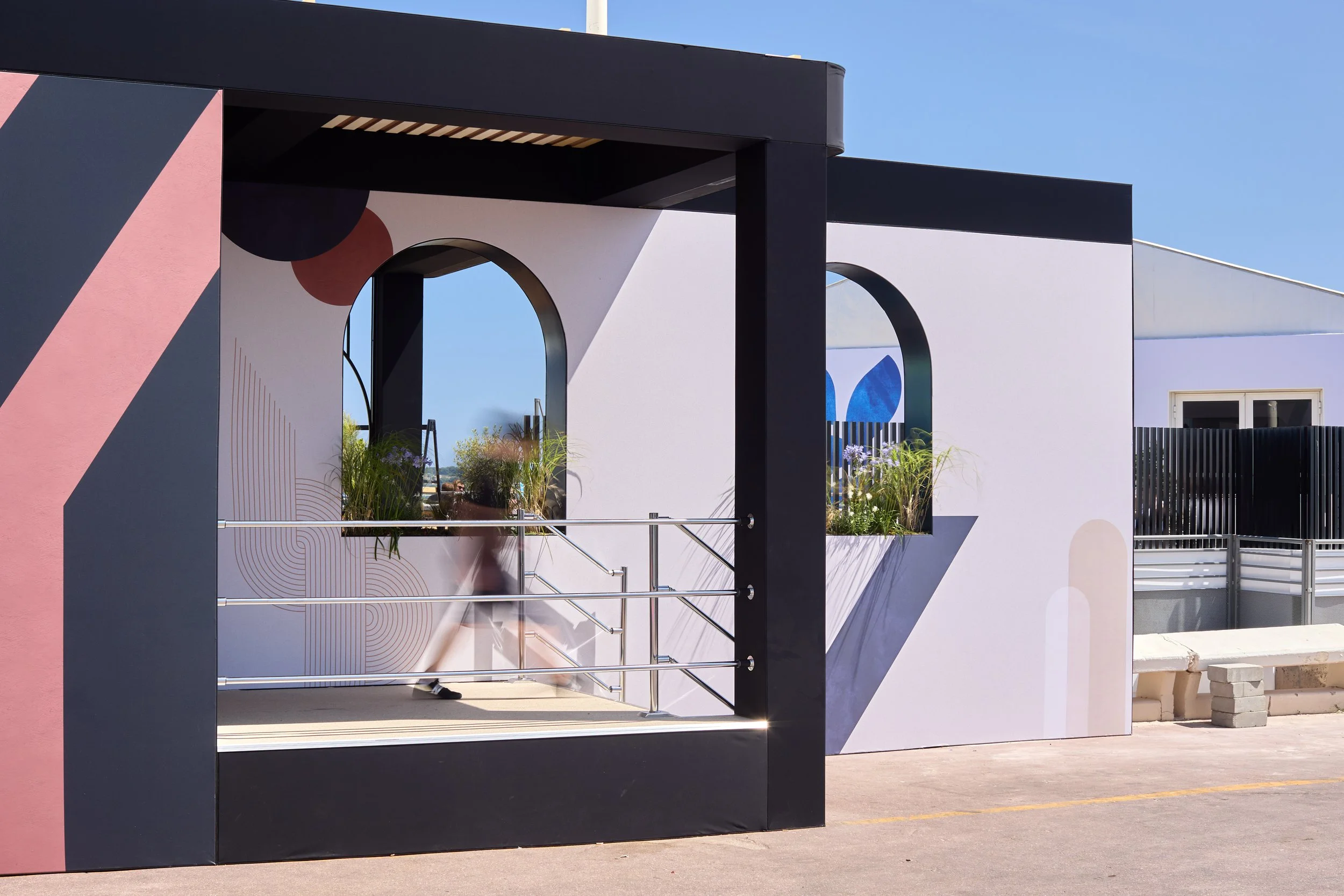 Modern building exterior with geometric mural, featuring an arched window with plants and a person walking by on a sunny day.