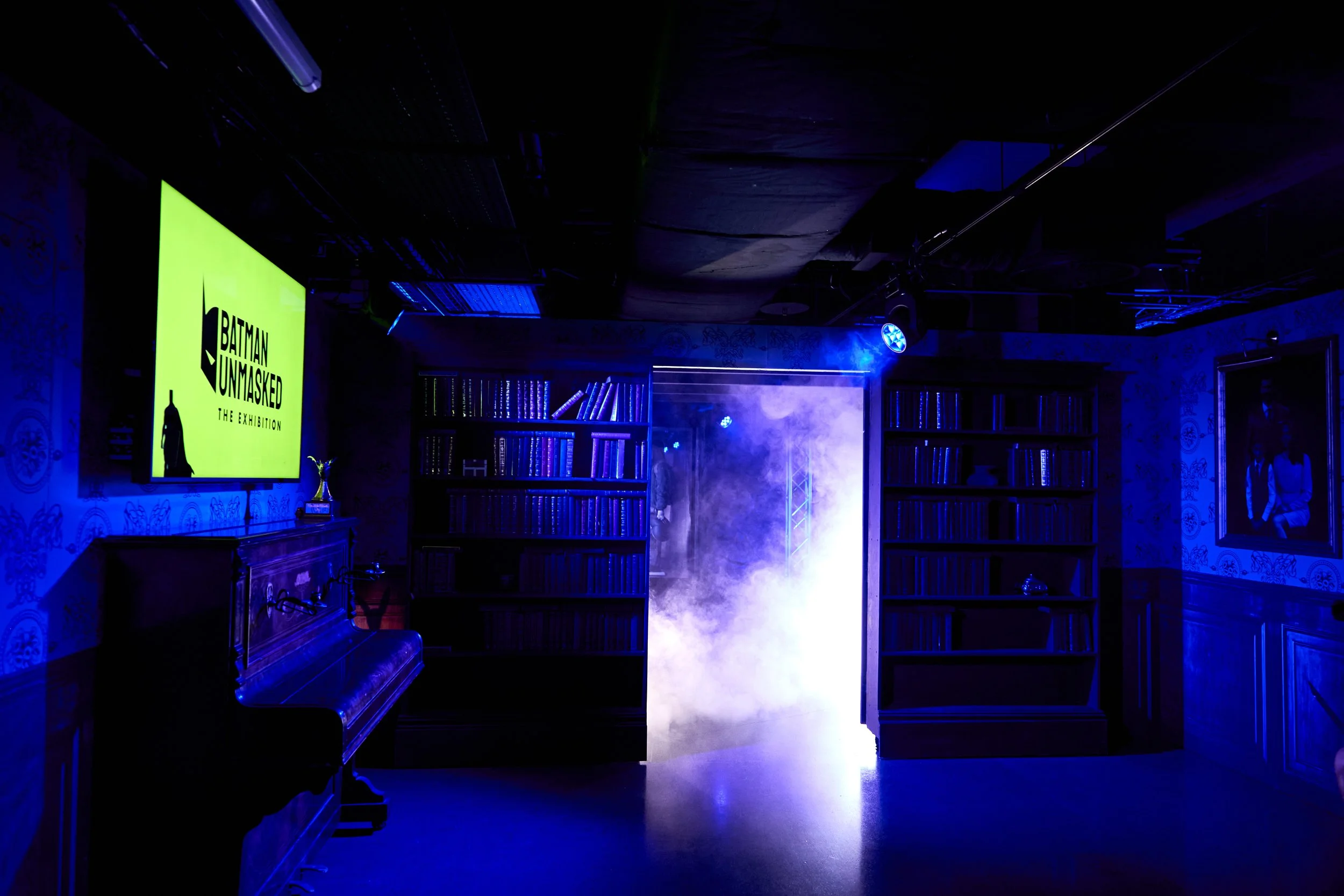 An indoor scene with a black piano on the left, a TV screen displaying 'BATMAN UNMASKED THE EXHIBITION' on the wall, bookshelves filled with books on both sides, a smoky fog in the center, and blue lighting creating a mysterious atmosphere.